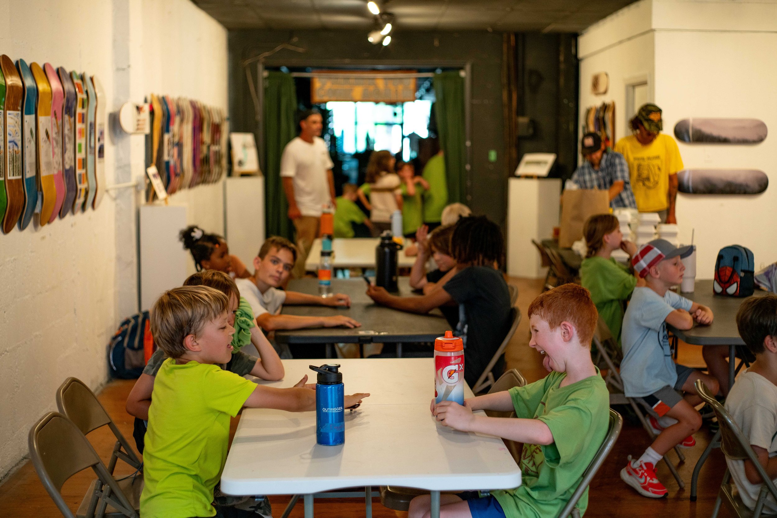 Children sitting at tables in an indoor skate-themed space, with skateboards displayed on the wall, and some adults and children in the background.