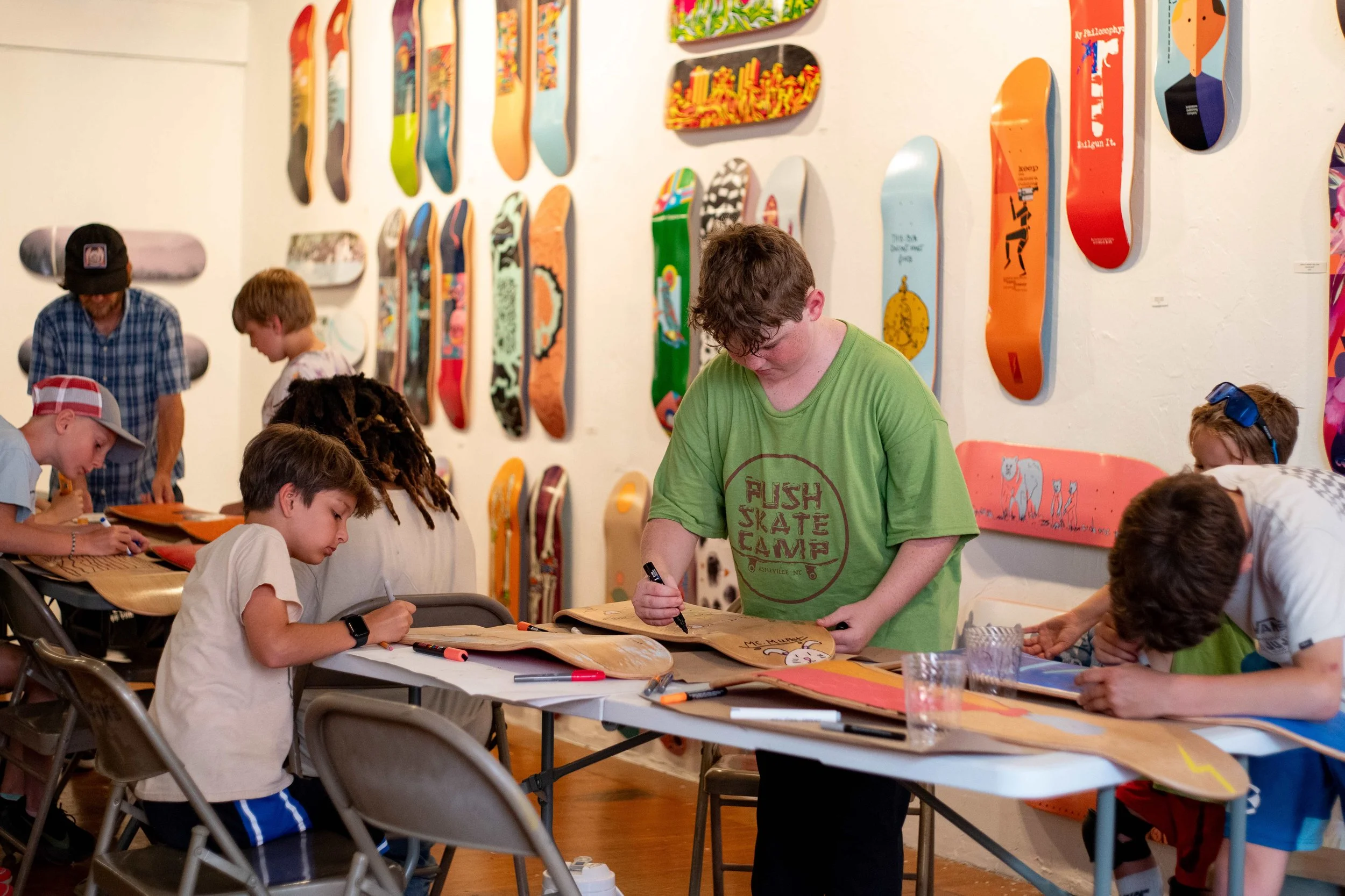 Children and adults at a skateboarding-themed art activity, drawing on skateboard decks, with skateboard art on the wall in the background.