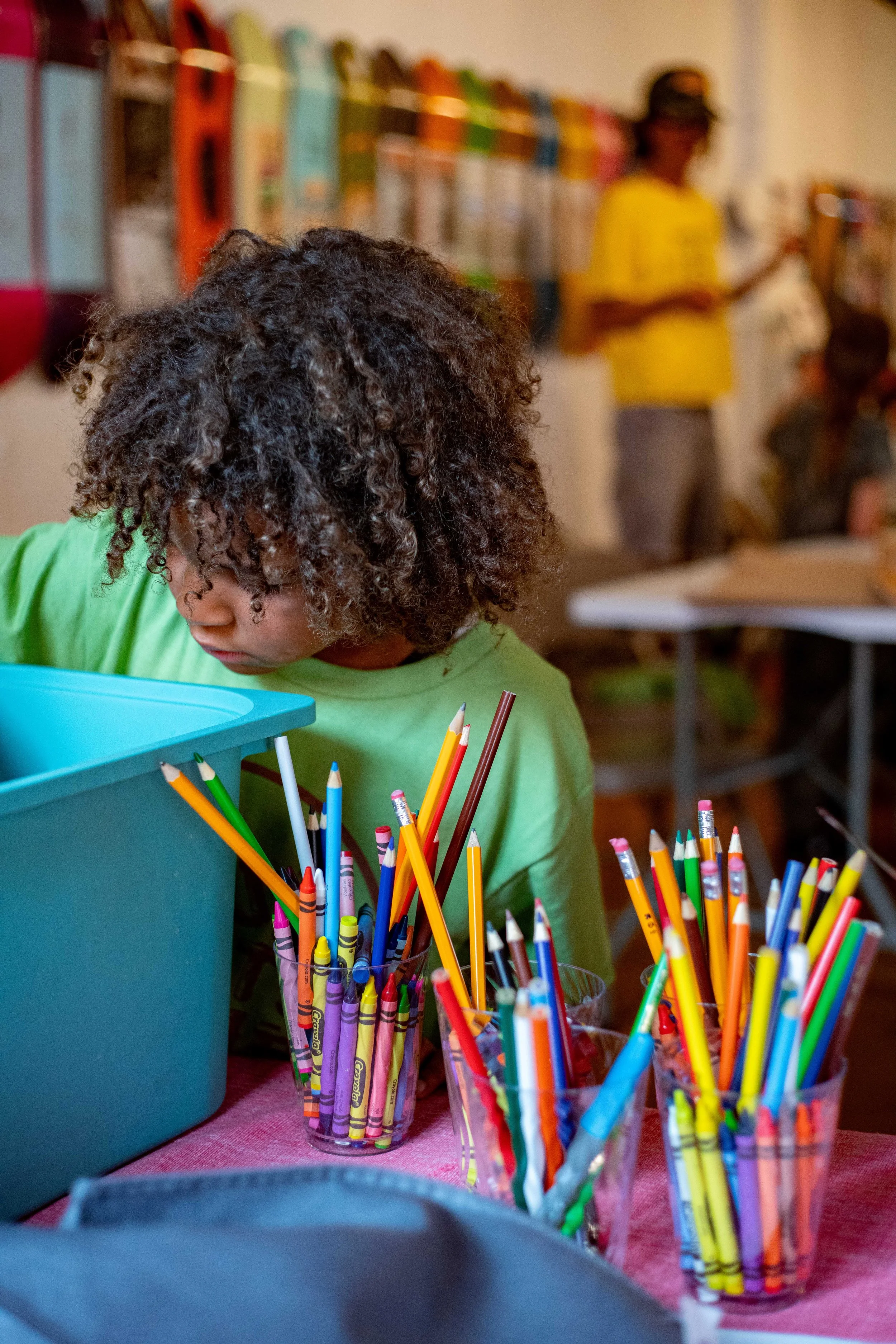 A young girl with curly hair in a green shirt organizing colorful pencils in cups on a table in a classroom.