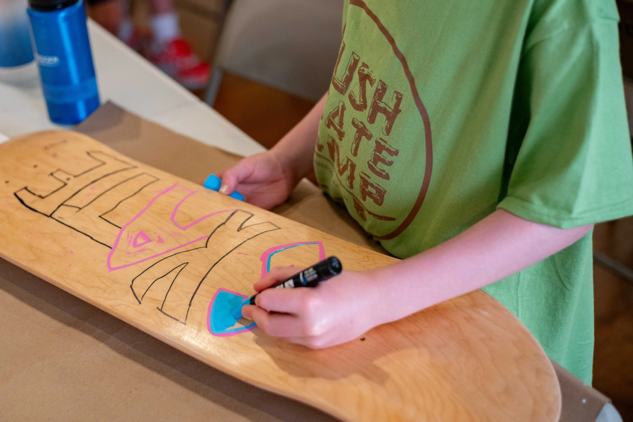 A child in a green shirt drawing colorful shapes and writing on a skateboard with a black marker.