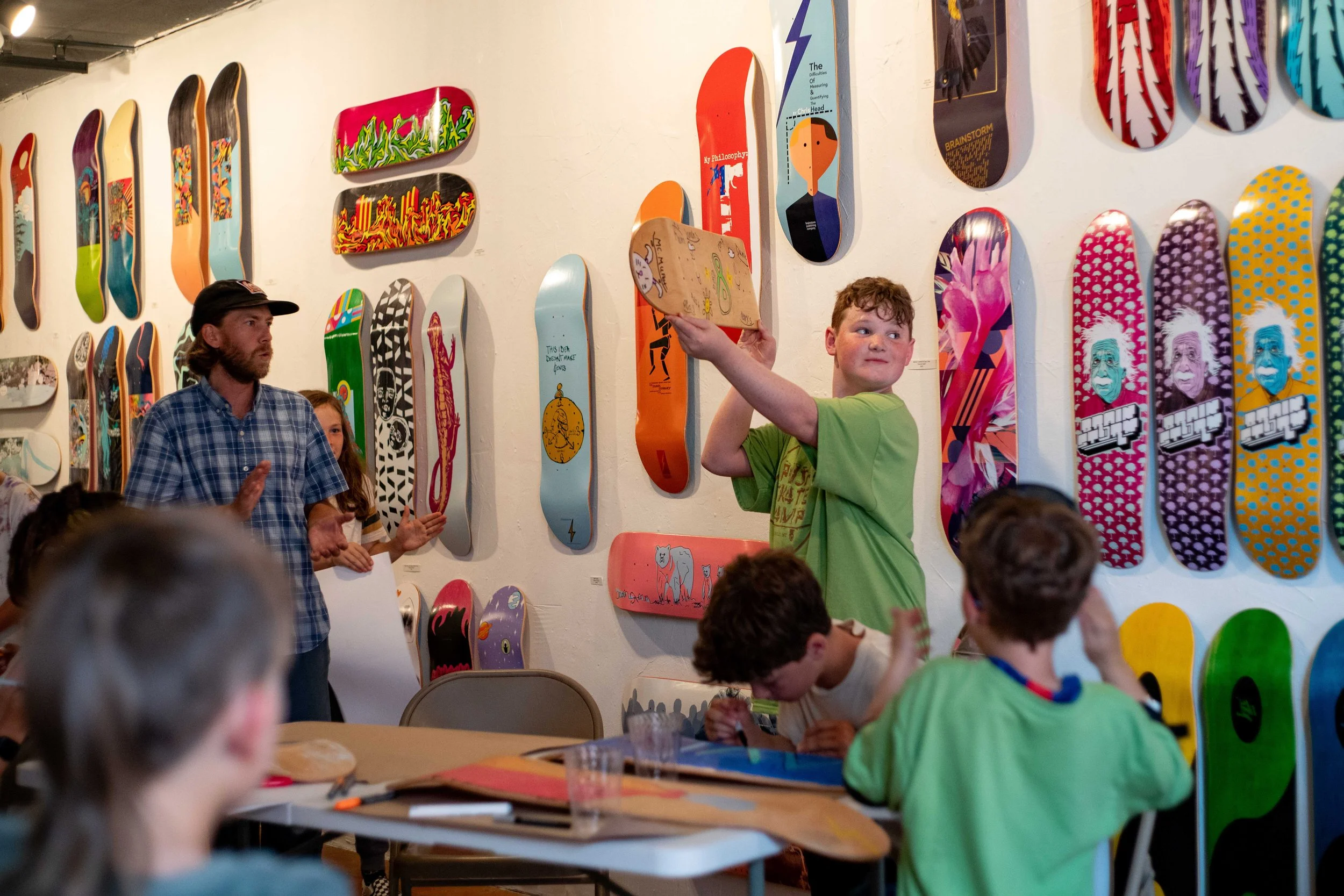 Children and an adult in an art gallery with skateboards hanging on the wall, some children are sitting at a table, and one boy stands holding a skateboard.