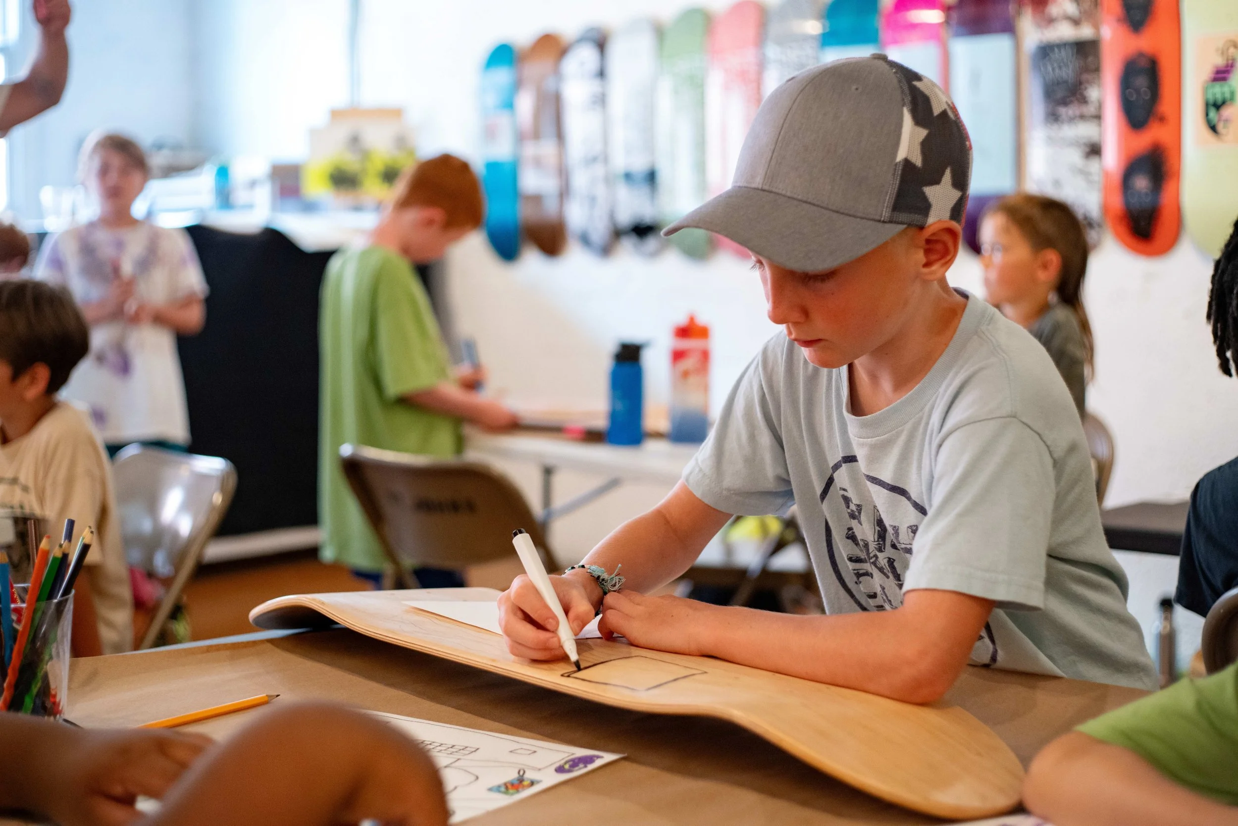 A young boy wearing a gray cap and a gray t-shirt drawing or writing with a black marker on a skateboard in a classroom with other children in the background.