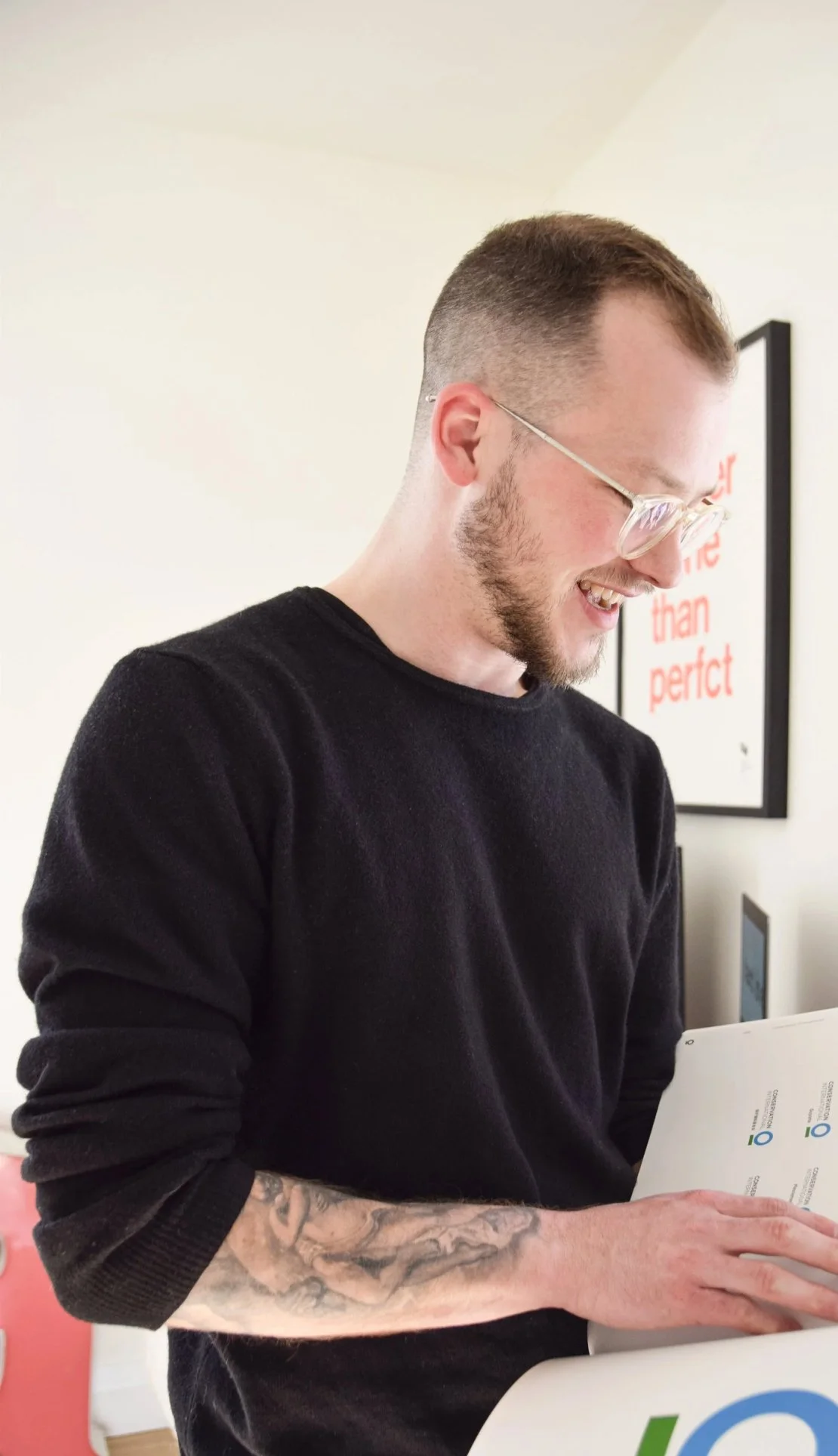 A smiling man with glasses and a tattooed arm wearing a black sweater, looking at a piece of paper or a document.