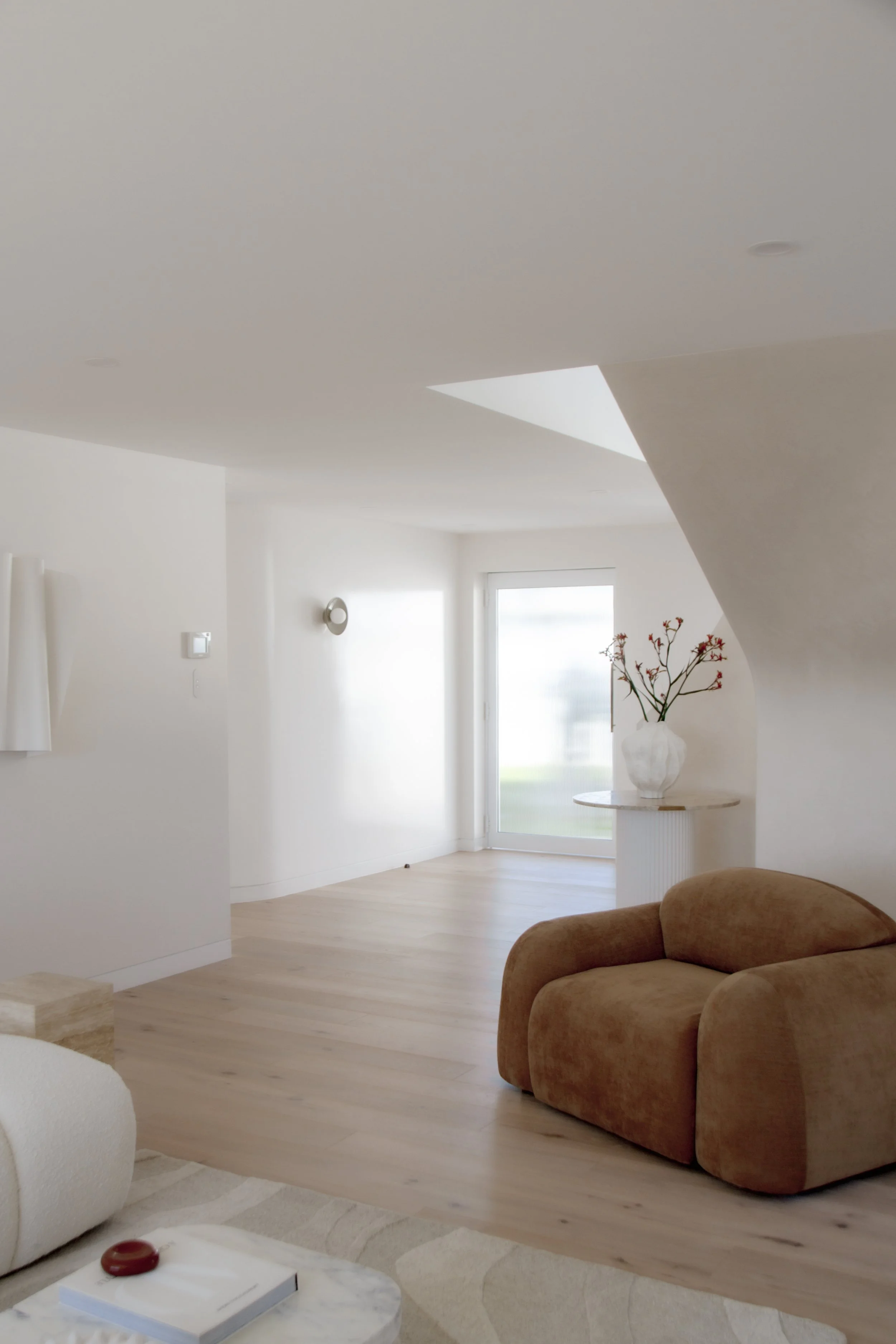 A minimalist living room with a beige armchair, a white ottoman, and a white vase with branches on a stand near a large window, featuring light wooden flooring and white walls.