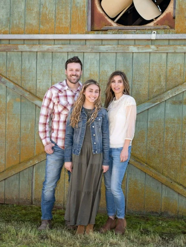 Three people standing outdoors in front of a green barn door, smiling at the camera. A man on the left wearing a plaid shirt and jeans, a young woman in the middle wearing a denim jacket and dress, and a woman on the right in a white blouse and jeans