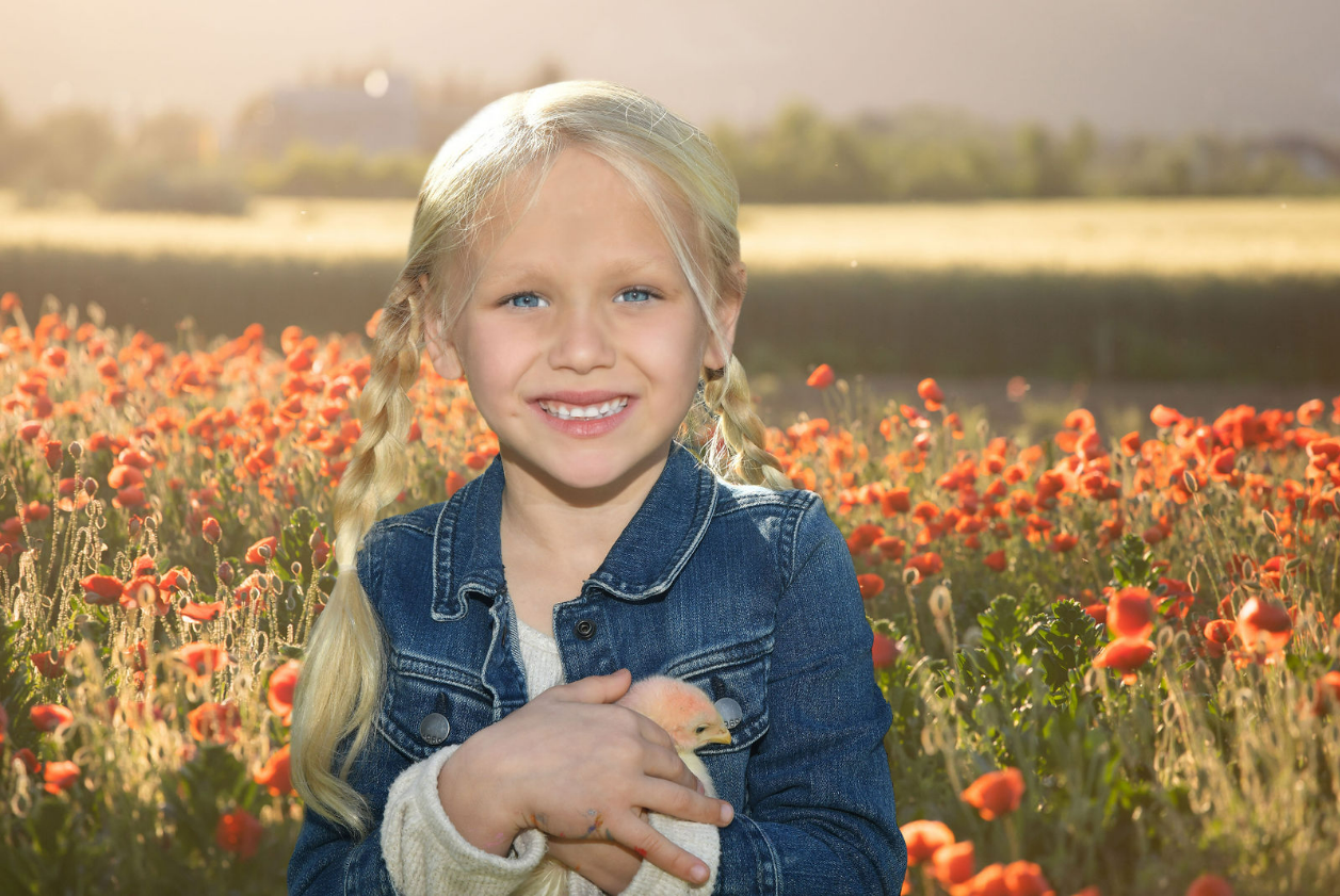 A young girl with blonde hair in braids, wearing a denim jacket, holding a small yellow chick, standing in a field of orange flowers with a blurred body of water and trees in the background on a sunny day.