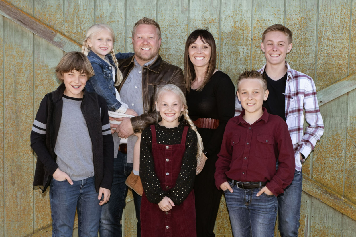 Family portrait of nine people including two adults and seven children posing together in front of a rustic wooden wall.