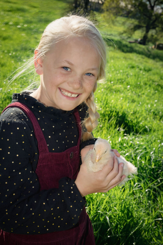 Young girl with blonde hair and freckles smiling outdoors in a grassy field, holding a fluffy white duckling.