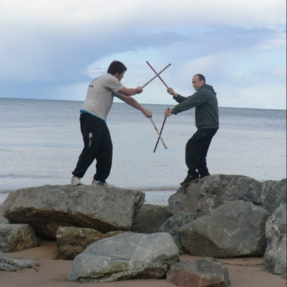 Two men on rocks at the beach engaging in a mock sword fight with sticks, with the ocean and cloudy sky in the background.