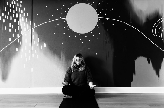 A woman sitting cross-legged on the floor in front of a black wall with white abstract and celestial artwork, including a large circle, lines, and dots.