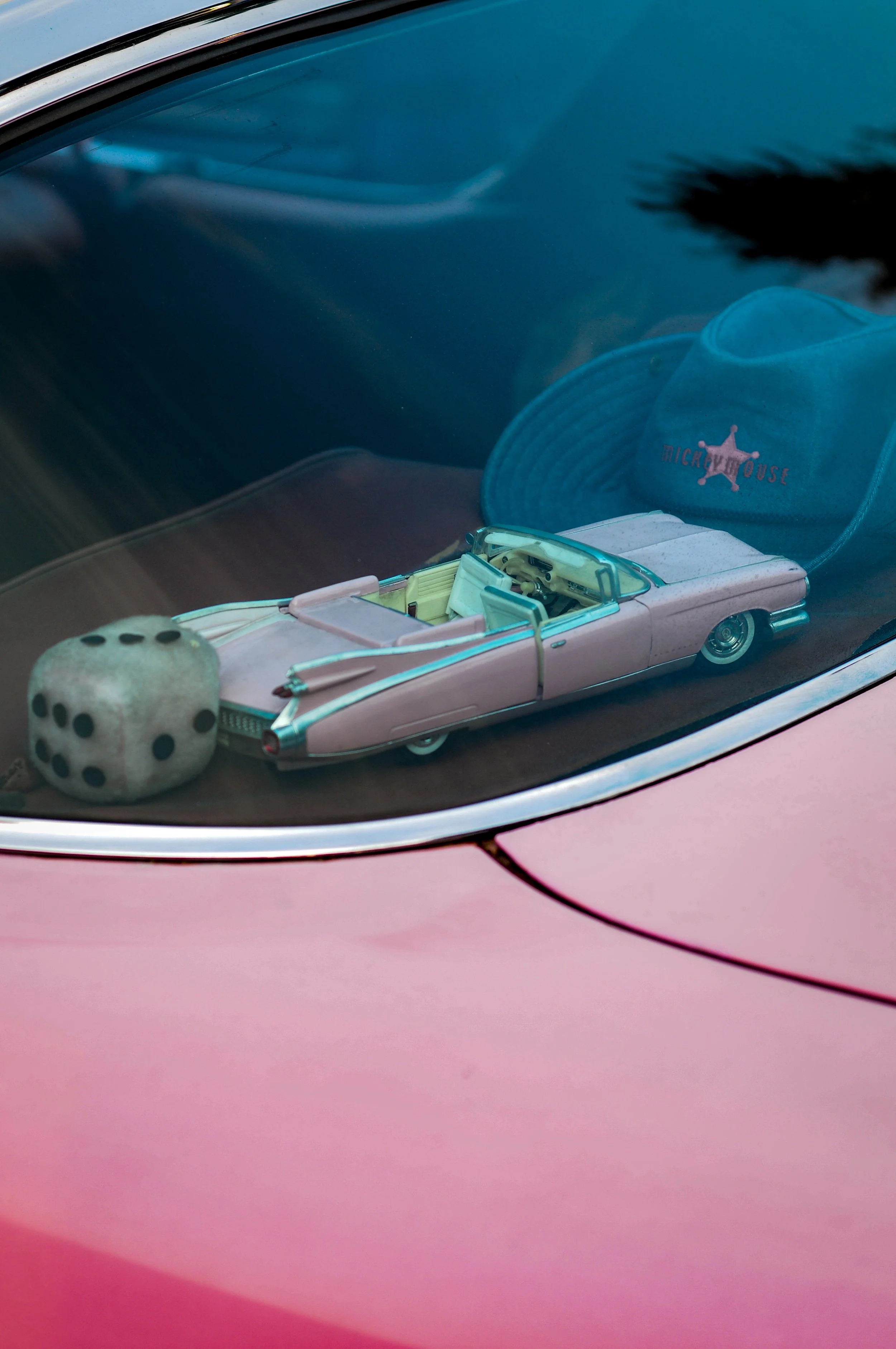 A pink toy car with a vintage style, a white fuzzy dice on the front, a light blue miniature cowboy hat with a pink star and the word 'MICKYHOUSE' on the inside, all placed on the dashboard of a vehicle with a dark interior and a pink exterior visible through the window.