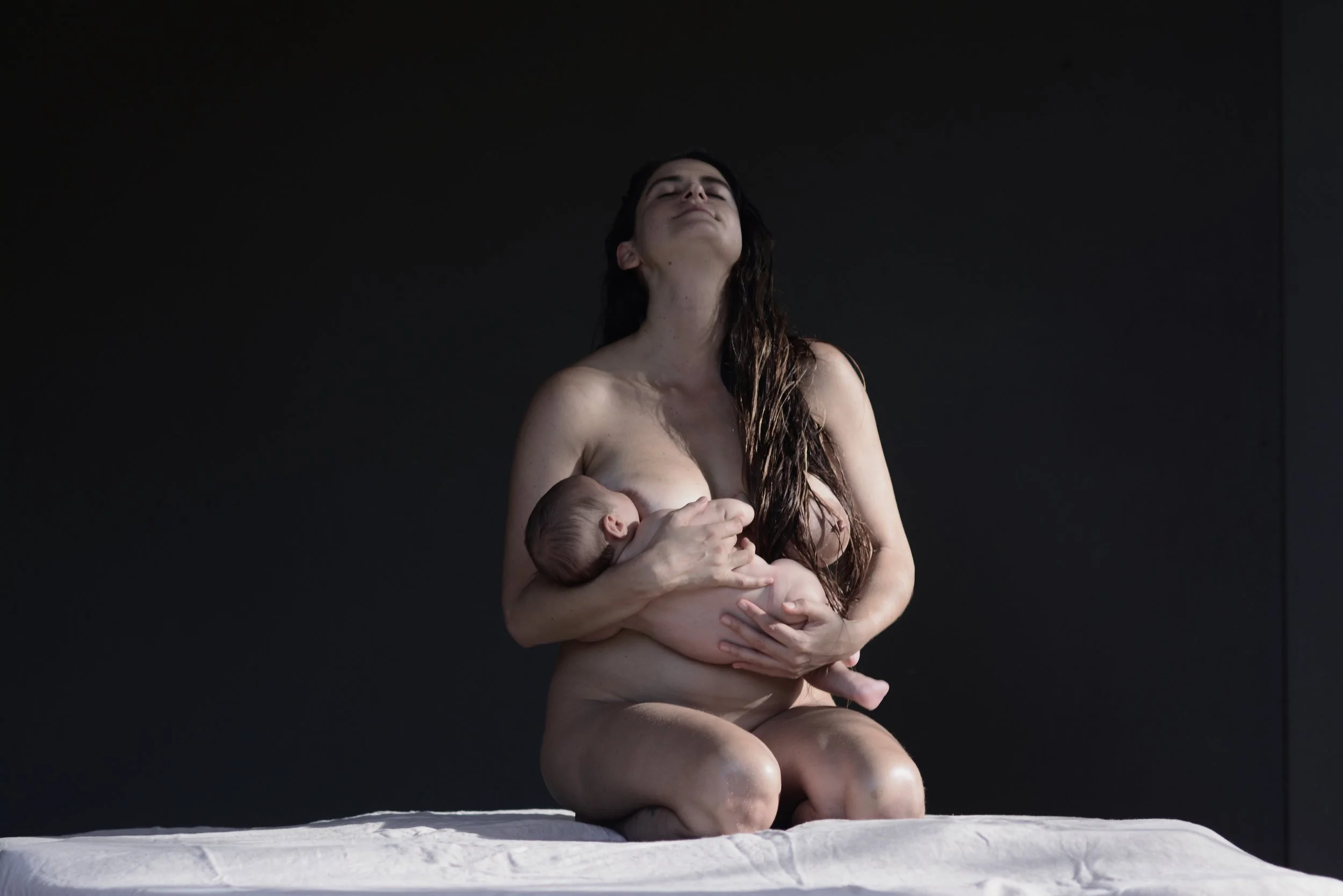 A woman with dark, wet hair holding a newborn baby on her lap against a dark background.