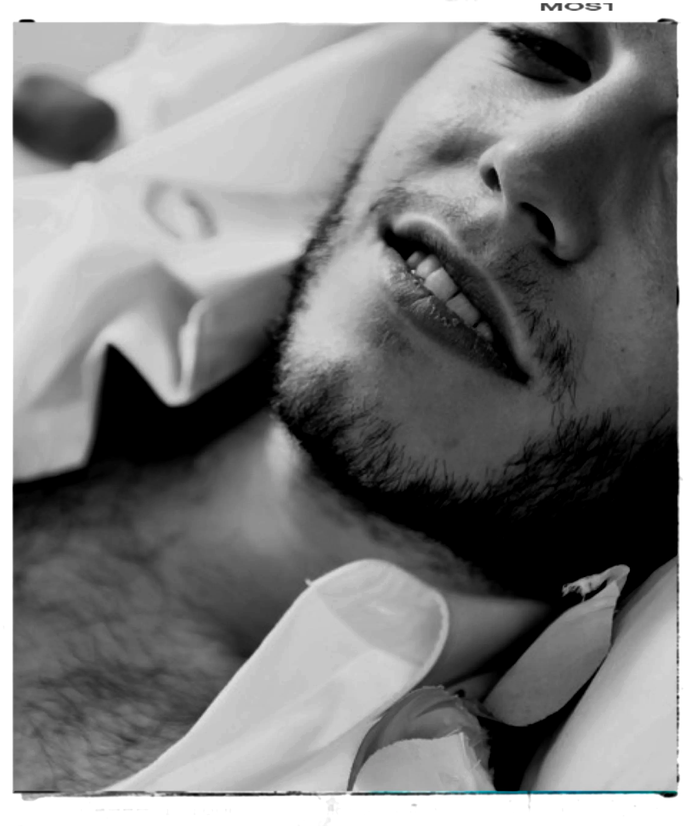 Close-up of a smiling man with facial hair lying down, with his head resting on a hand, in black and white.