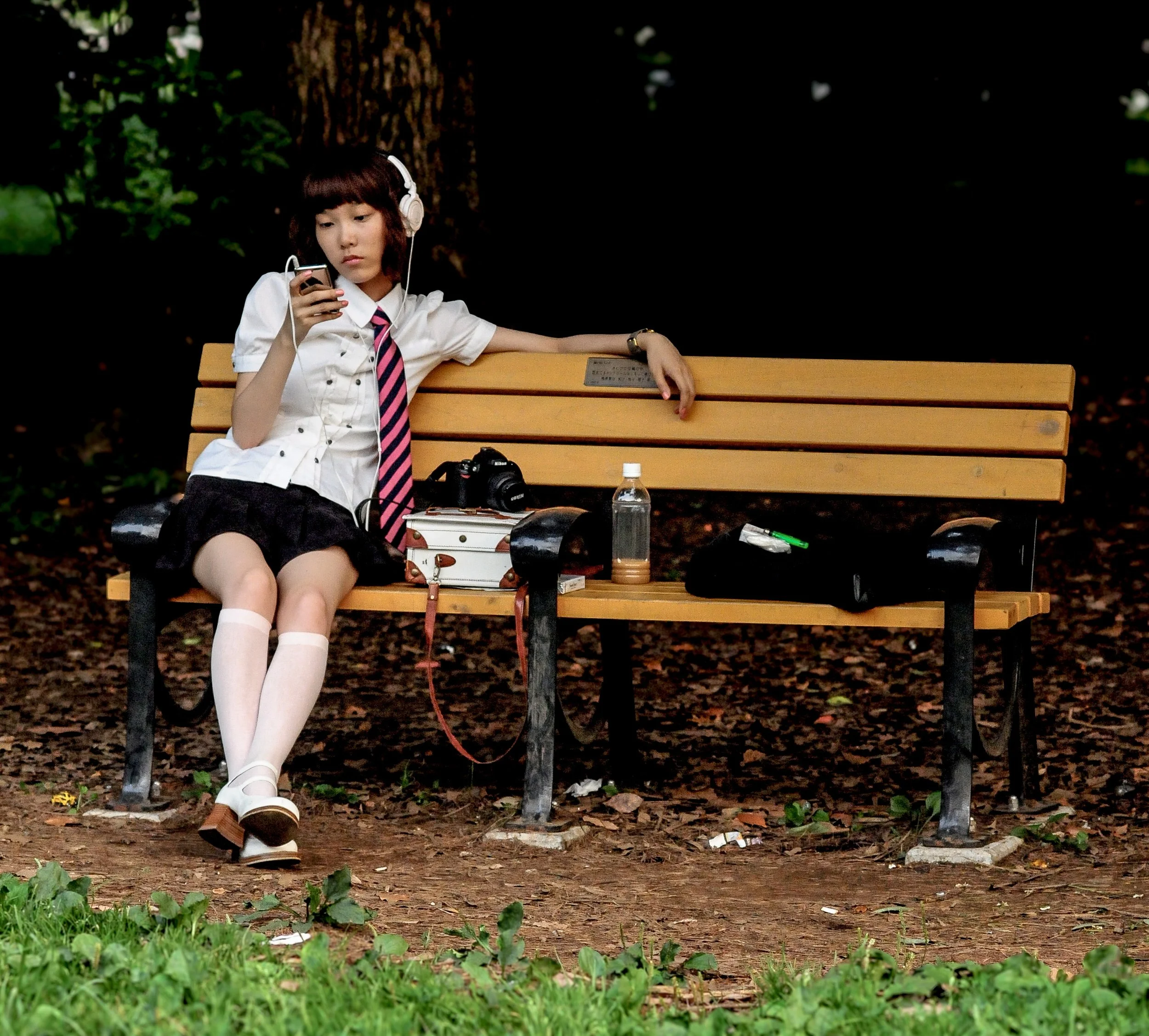 Young woman in school uniform sitting on a park bench, looking at her phone, with a camera and drink placed on her lap