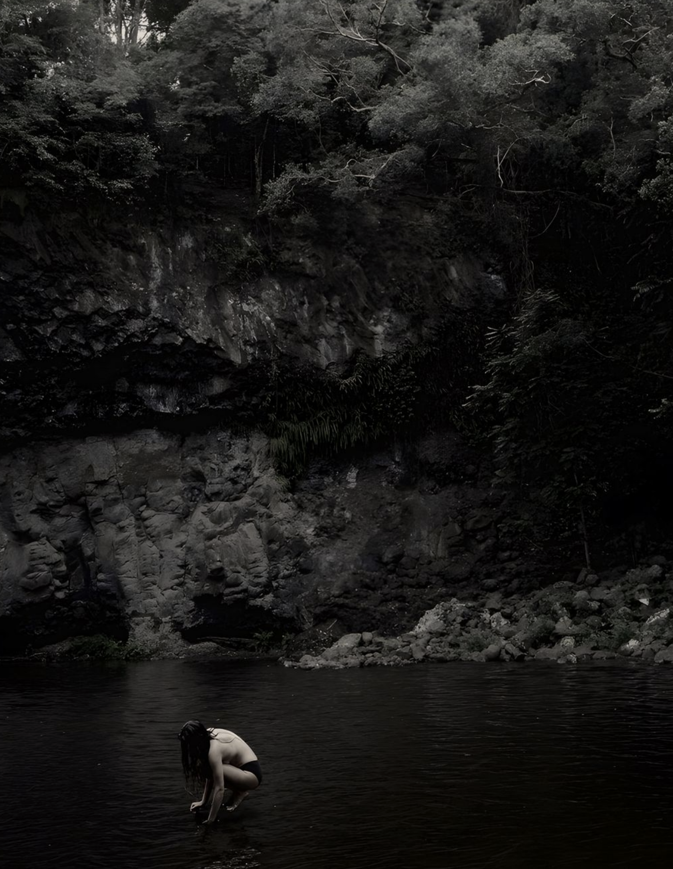 A person with long dark hair crouching in a dark river, surrounded by rocky cliffs and dense forest.