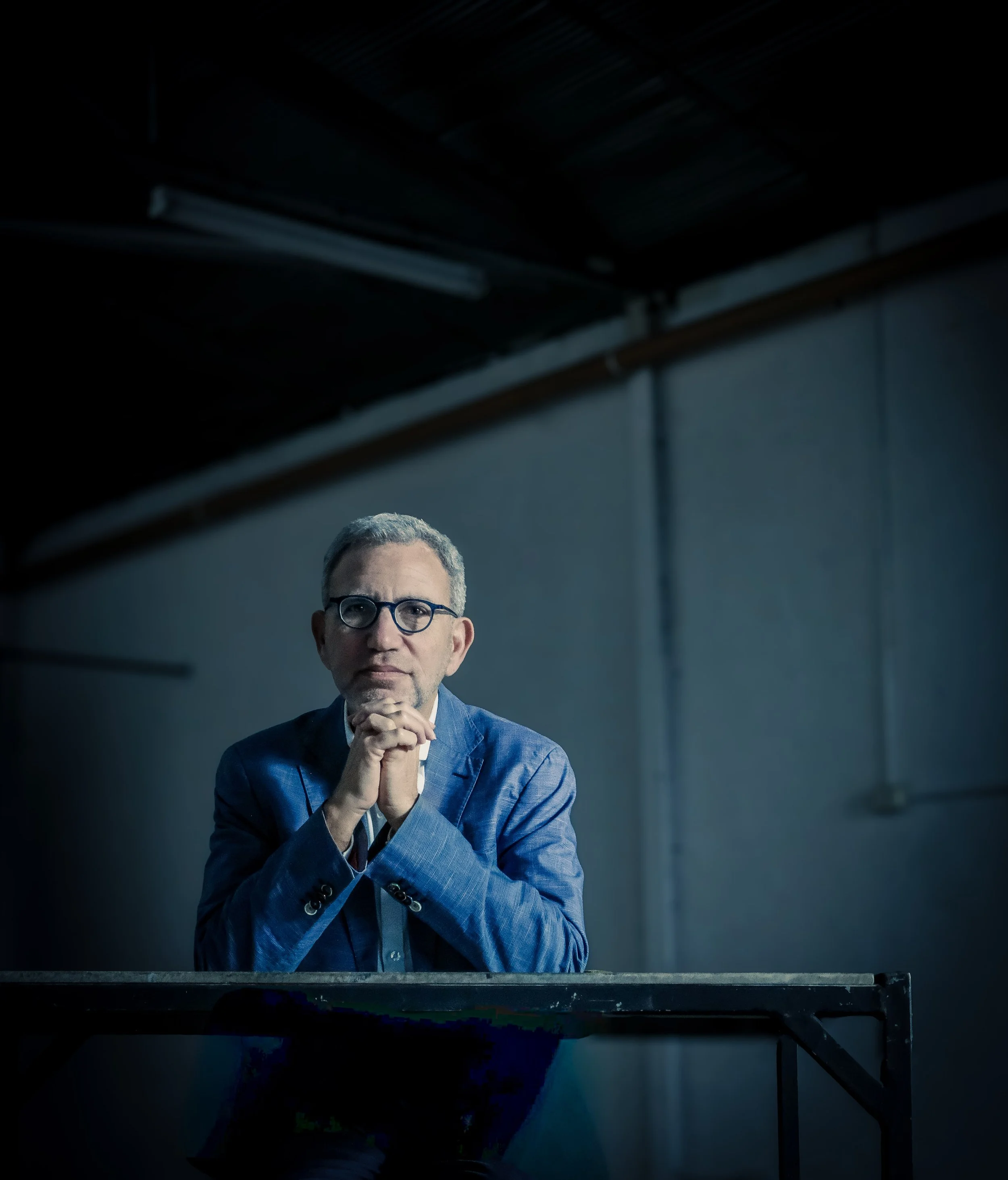 A man with glasses and gray hair wearing a blue blazer, sitting at a desk with his hands clasped together, in a dark studio setting.