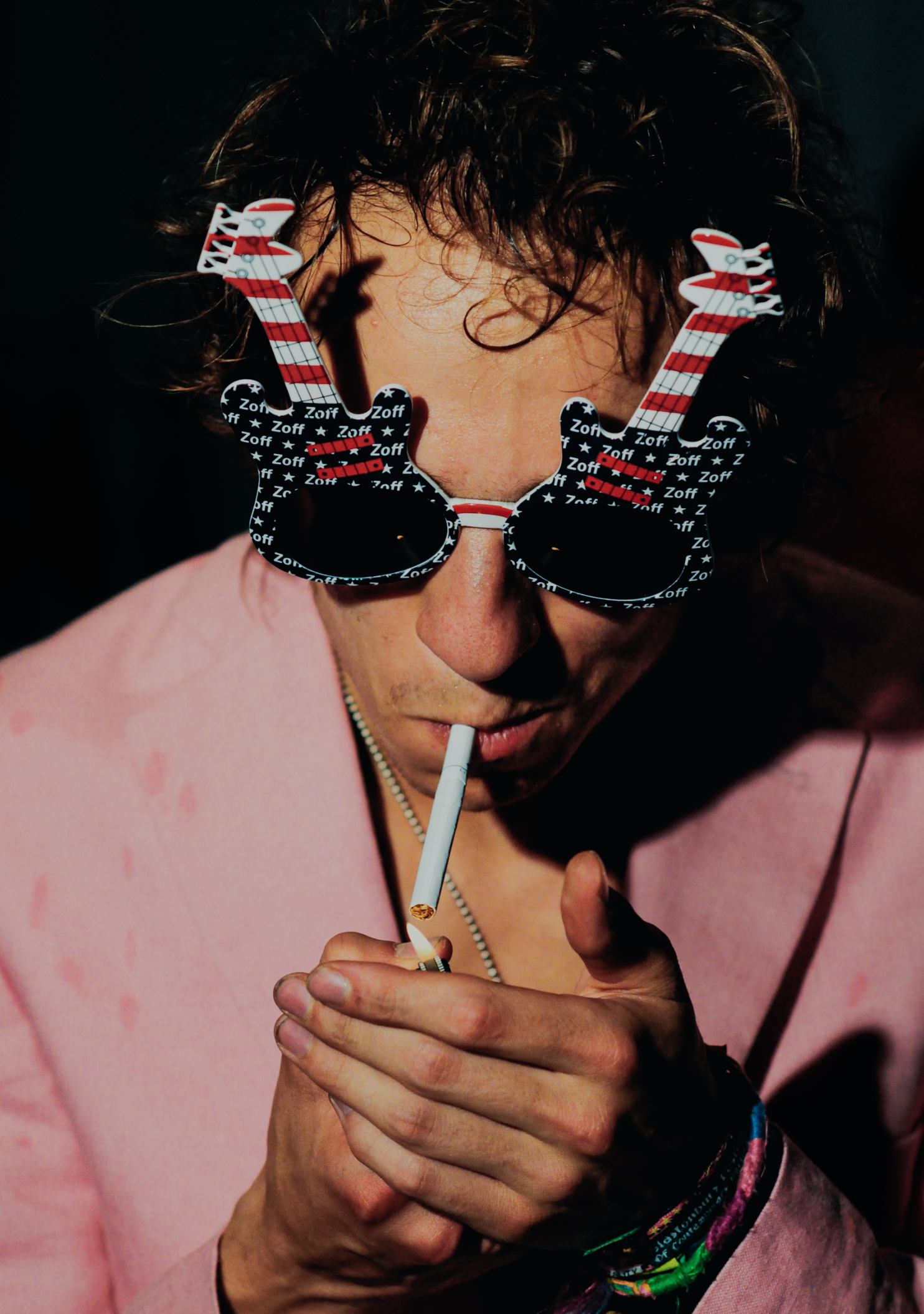 Person wearing novelty sunglasses with reindeer antlers design, lighting a cigarette with a match, dressed in a pink shirt.