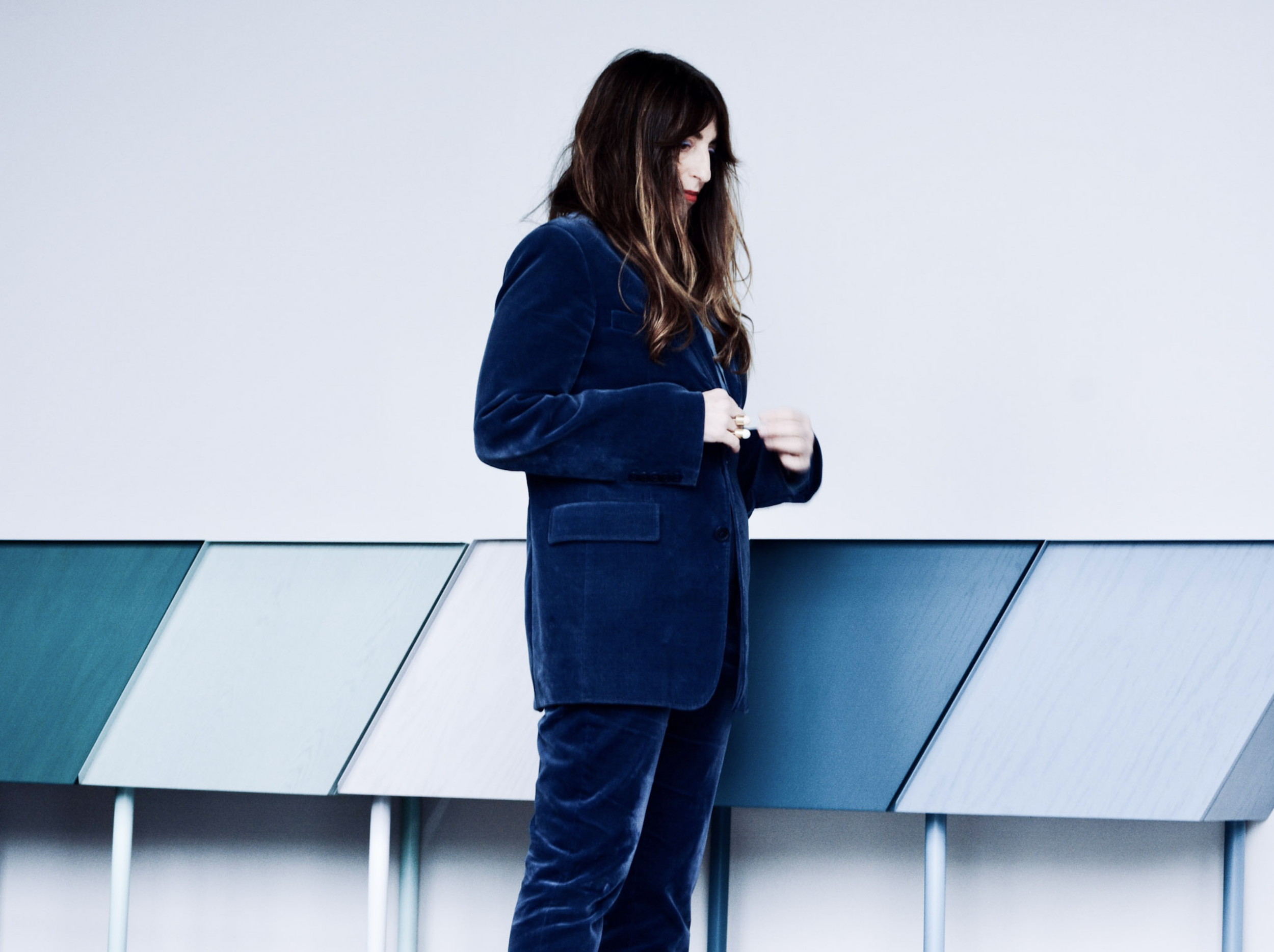 A woman with dark brown hair wearing a blue velvet blazer and matching pants standing against a plain wall and a modern, multi-colored cabinet.