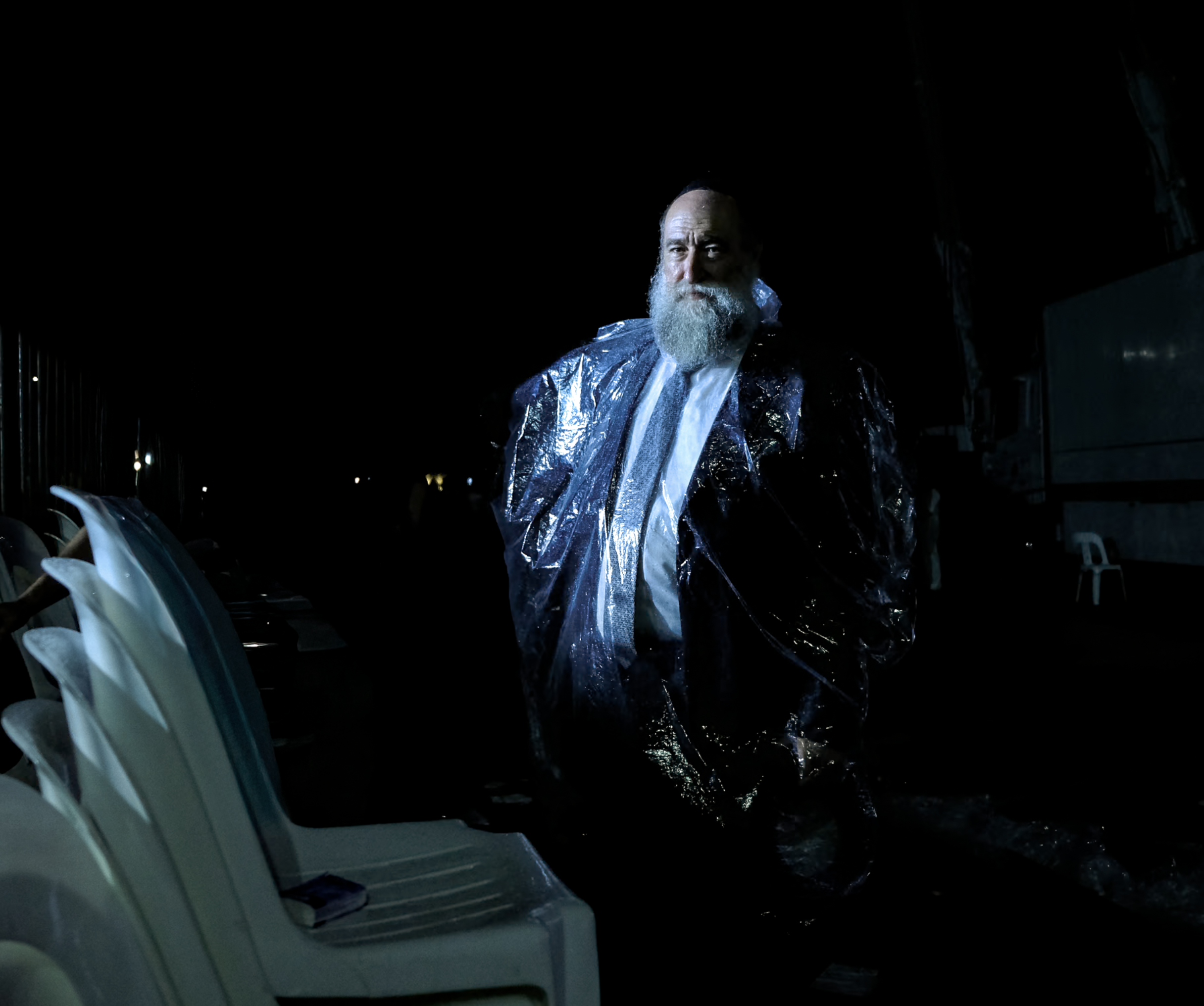 A man with a beard wearing a shiny, dark raincoat stands in the dark, illuminated slightly, with chairs in the background.