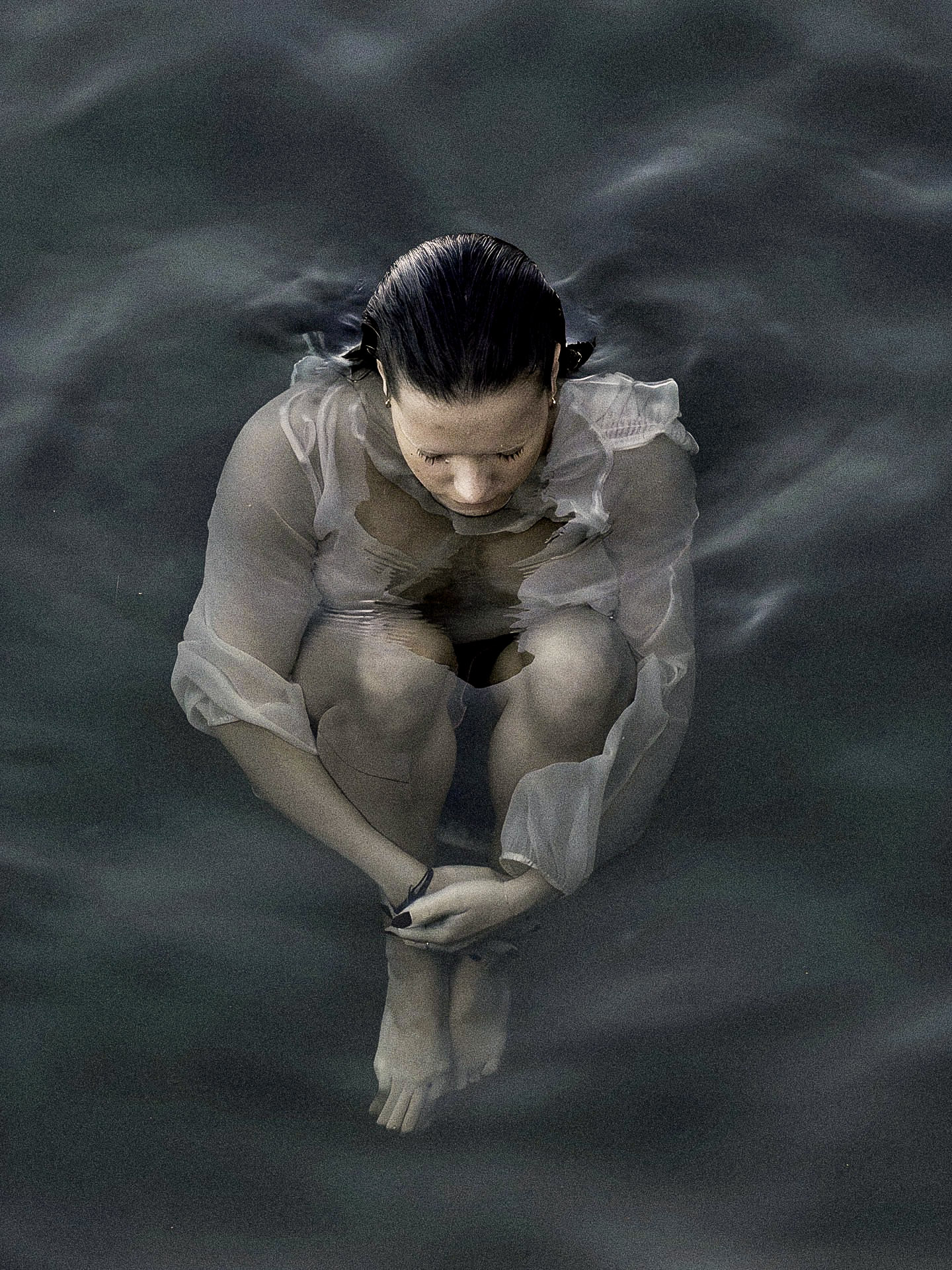 A person dressed in a white dress with long sleeves, sitting in water with closed eyes and head bowed, hair slicked back, surrounded by calm water.