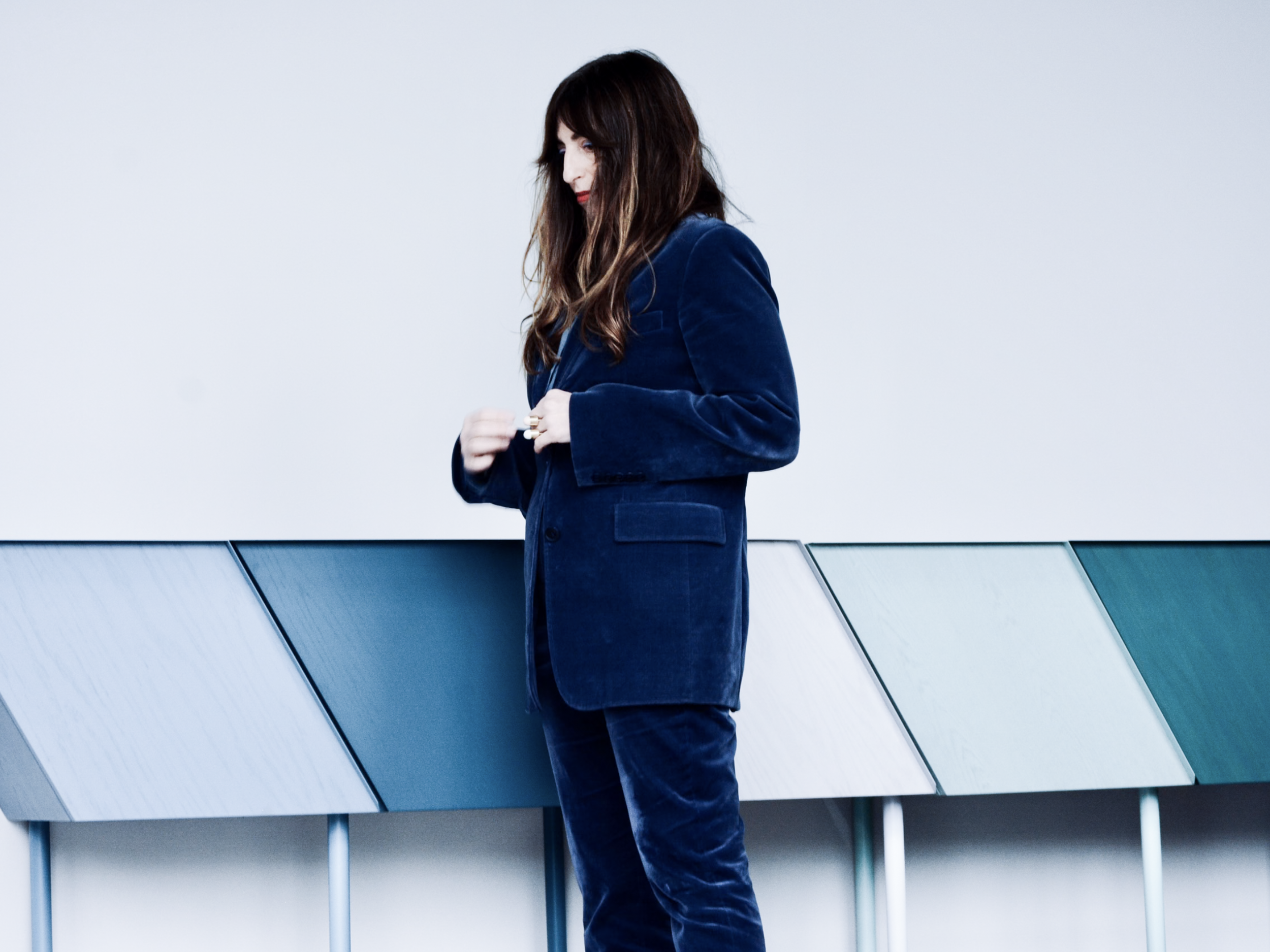 A woman in a navy blue velvet blazer and pants standing against a colorful panel wall, looking down with her hands adjusting her blazer.