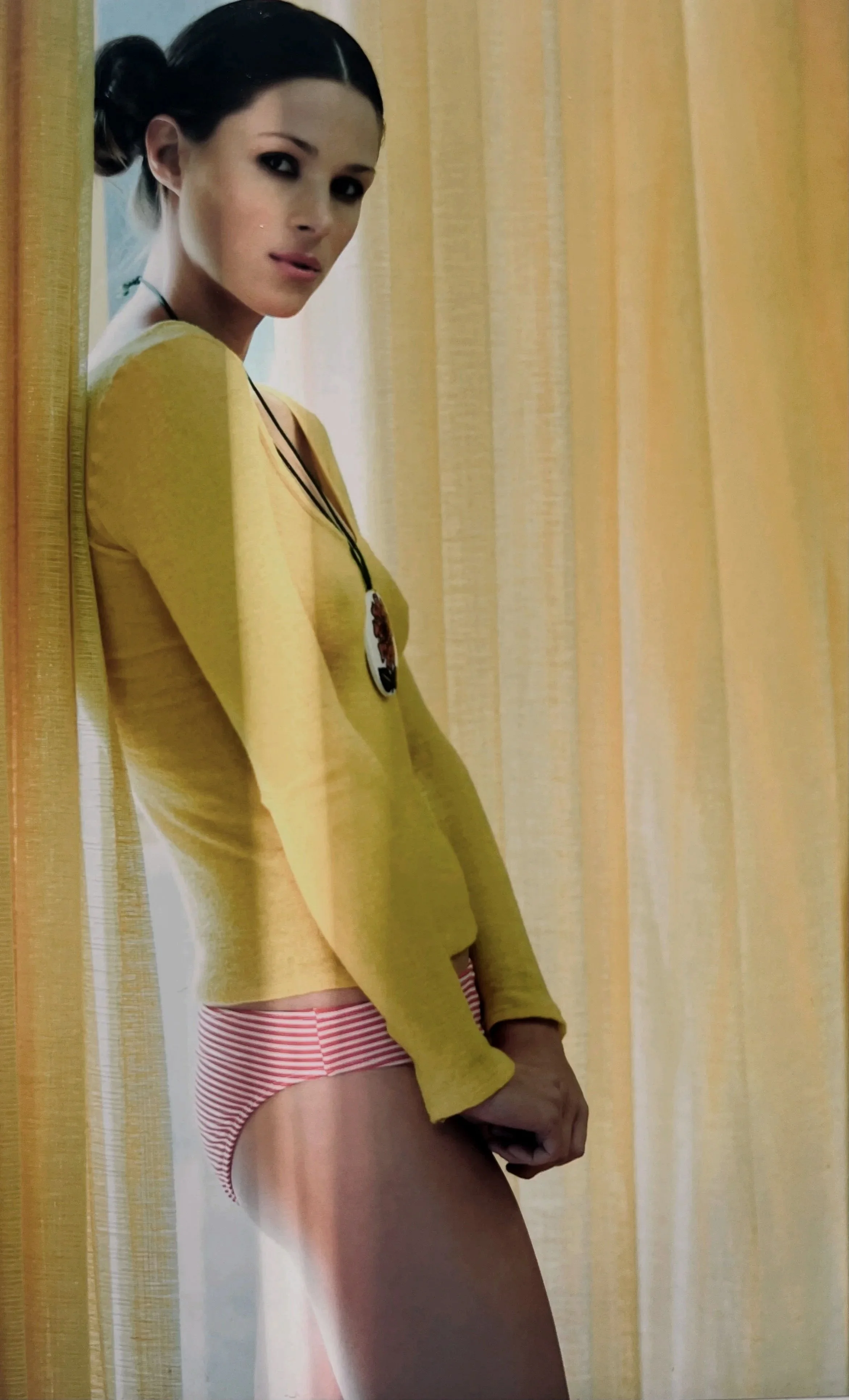 A woman with dark hair tied in a bun, wearing a yellow long-sleeve top with a pendant necklace and red-and-white striped underwear, standing by yellow curtains, looking at the camera.