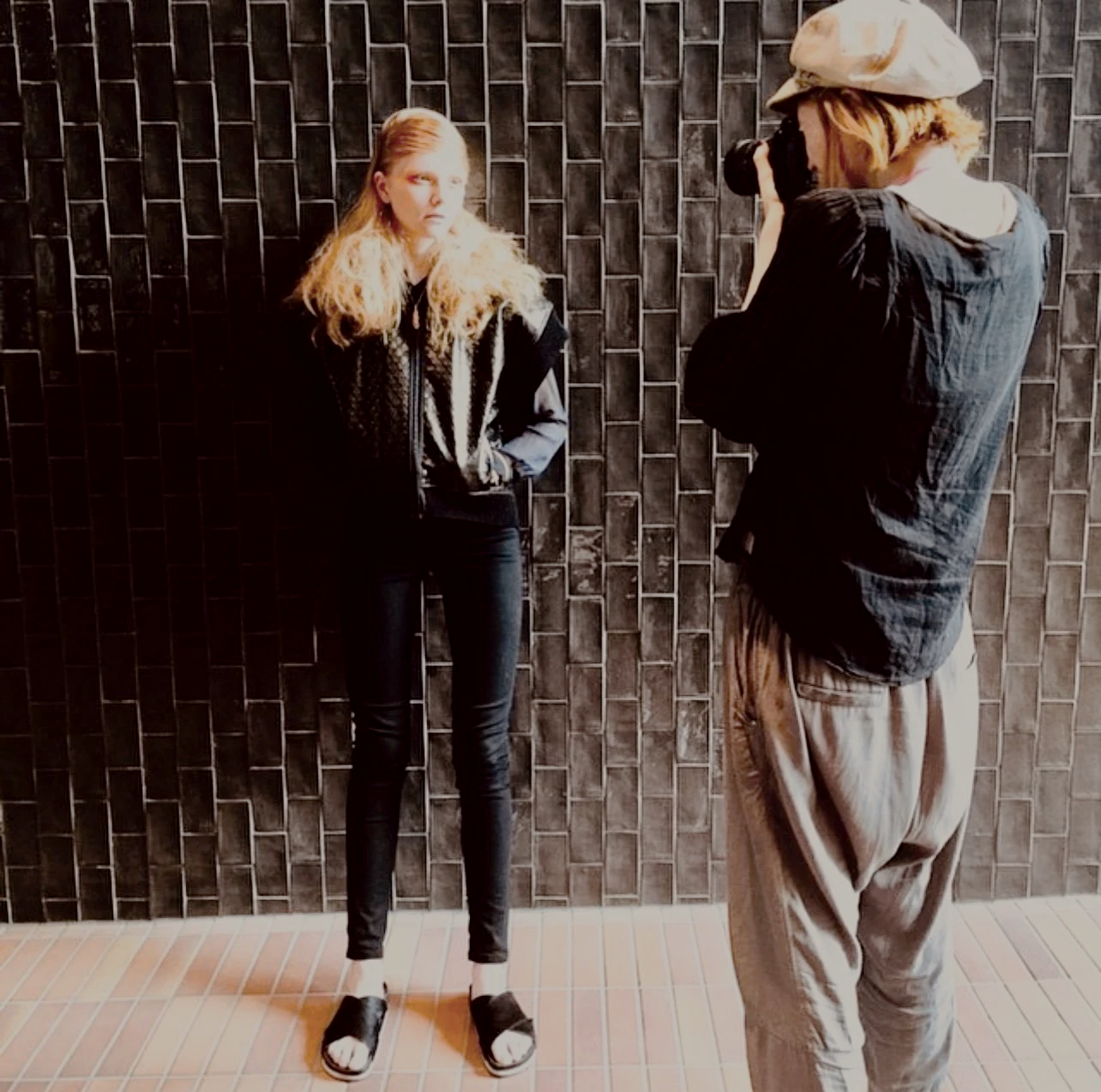 A woman with long blonde curly hair is posing for a photo in front of a dark tiled wall while a photographer captures the picture.