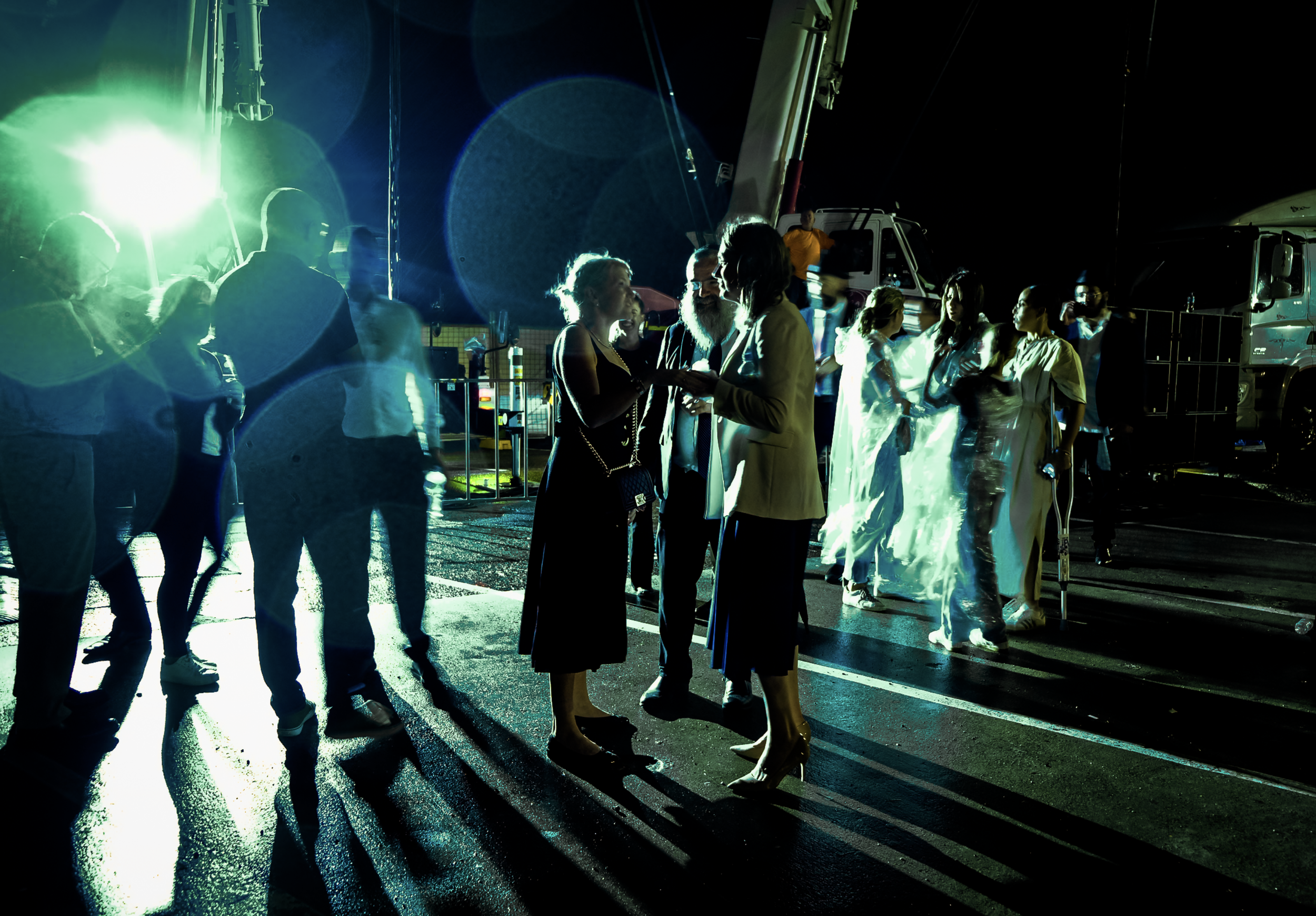 Bondi Chanukkah.. Group of people socializing outdoors at night, illuminated by bright green light, with large trucks and equipment in the background.