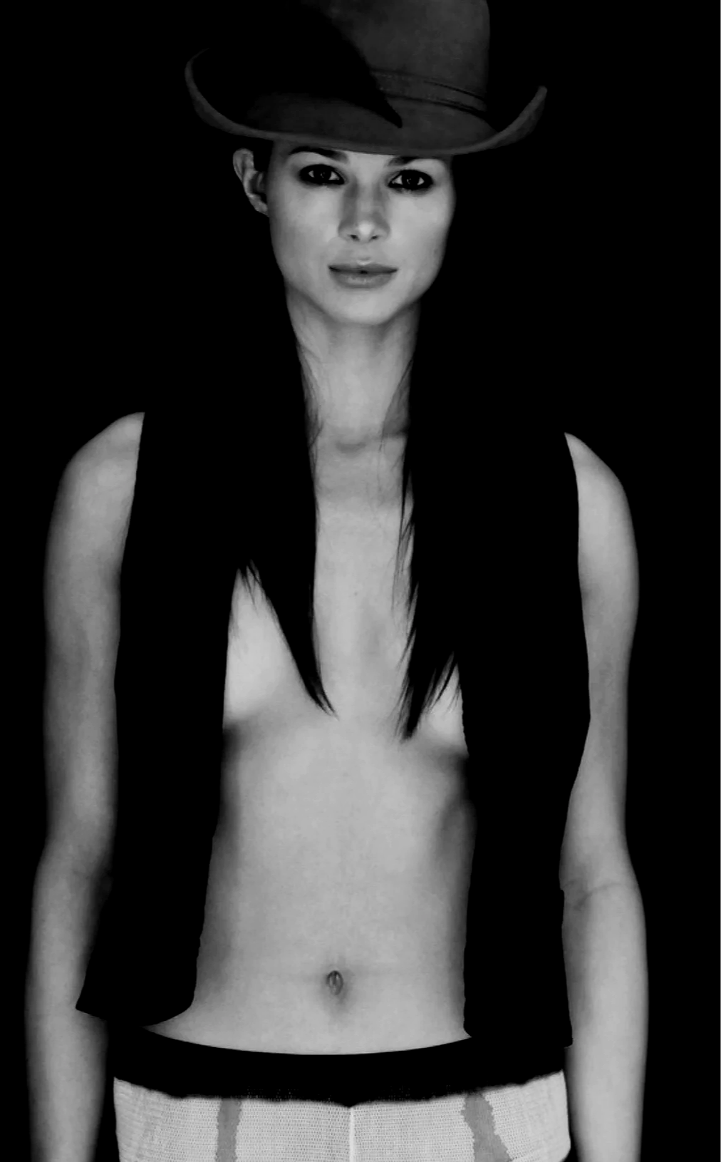 Black and white photo of a woman wearing a cowboy hat, with long dark hair, a sleeveless vest, and a navel piercing, against a dark background.