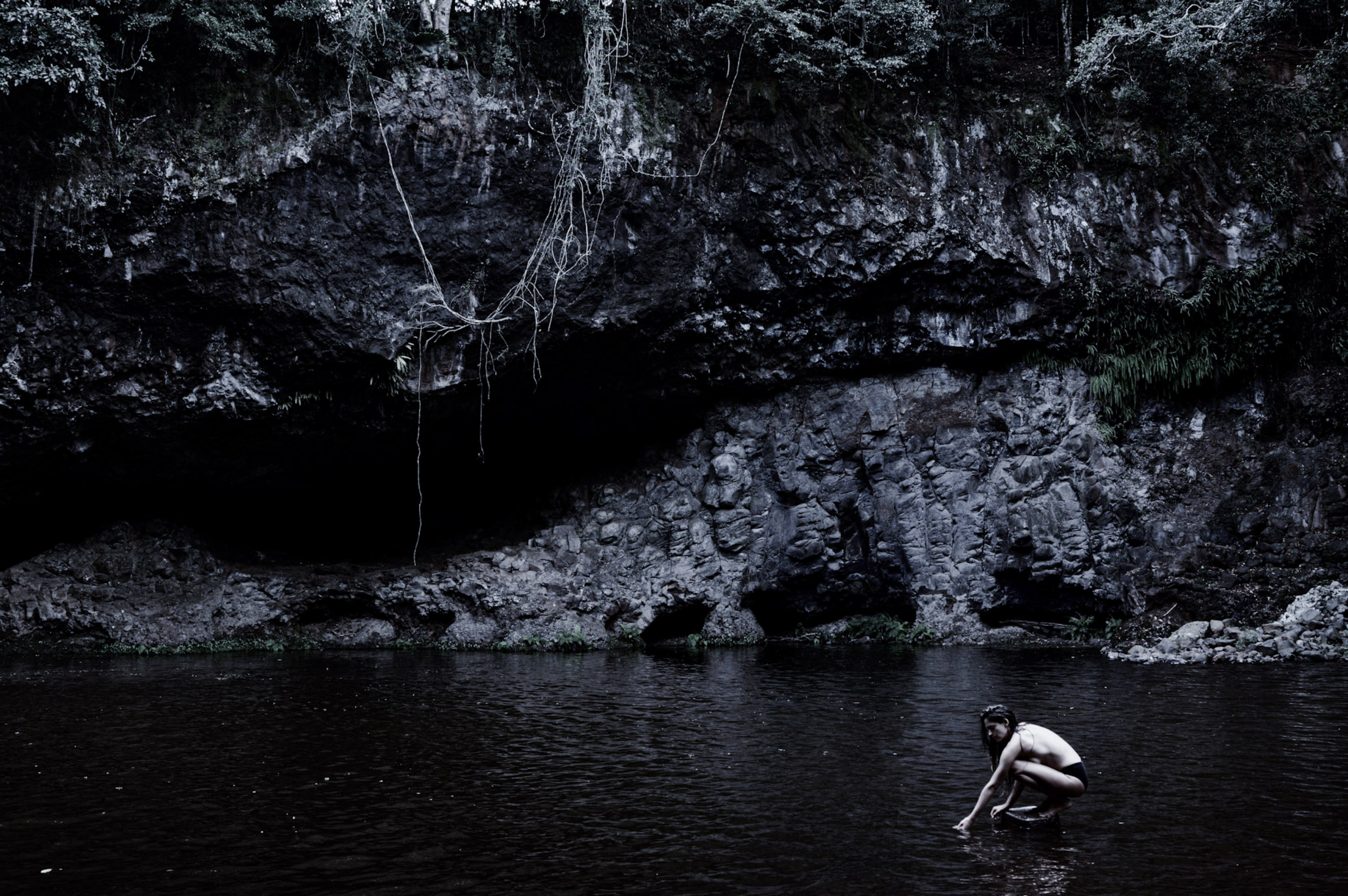 Person crouching in dark water with a rocky cave and foliage behind in a natural outdoor setting.