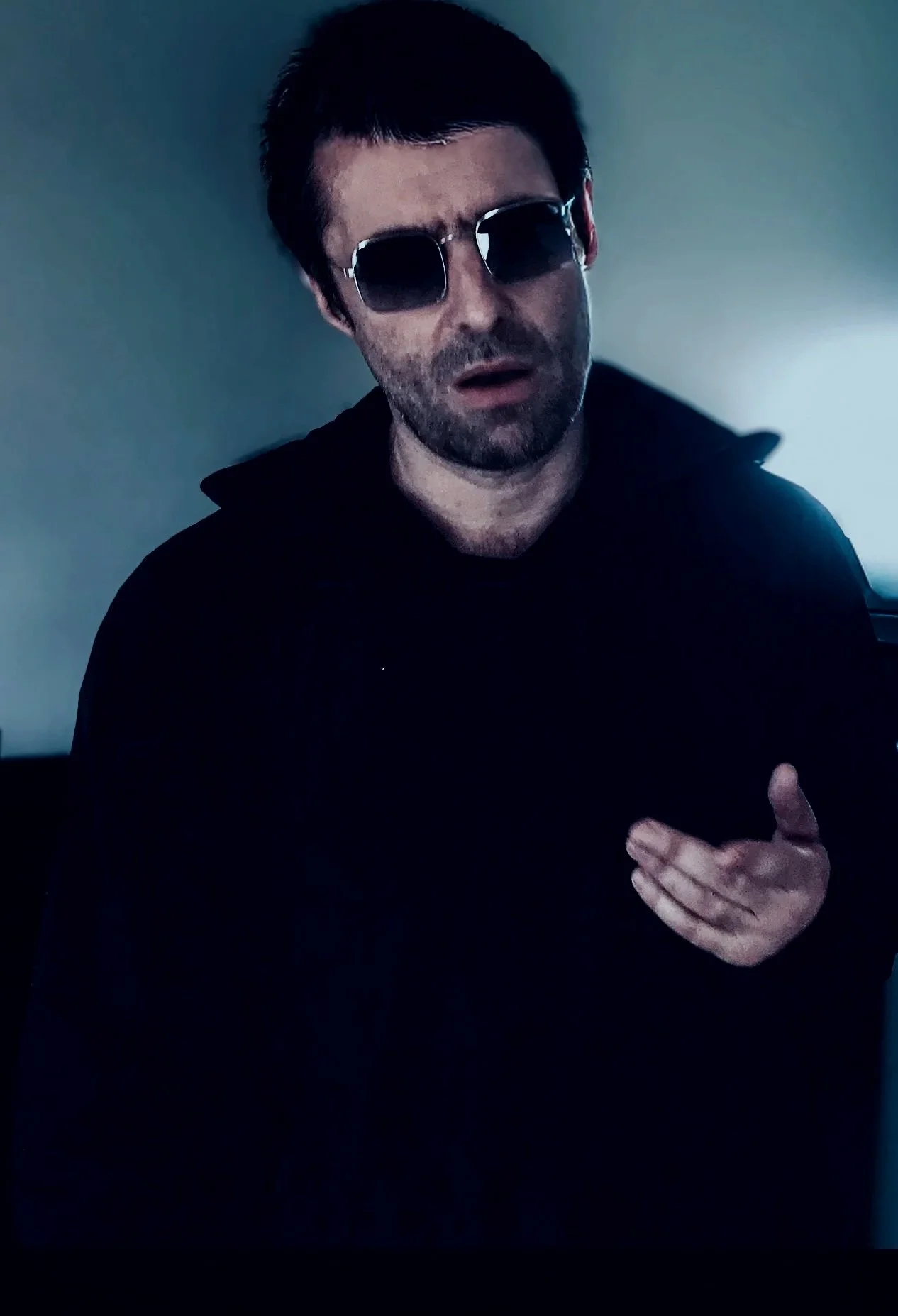 Primal Scream, Mani, Bobby Gillespie, Liam Gallagher, A man with dark hair, wearing dark sunglasses and a black jacket, gesturing with his right hand, in a dimly lit room.