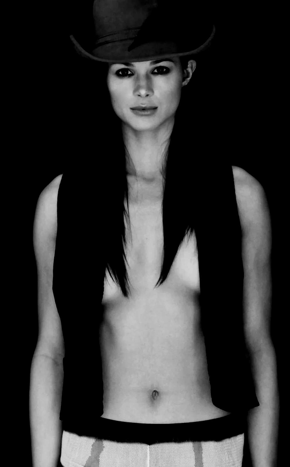 Black and white portrait of a woman wearing a wide-brimmed hat, with long dark hair, dark eye makeup, and a confident expression, topless with bare shoulders and a visible navel piercing.