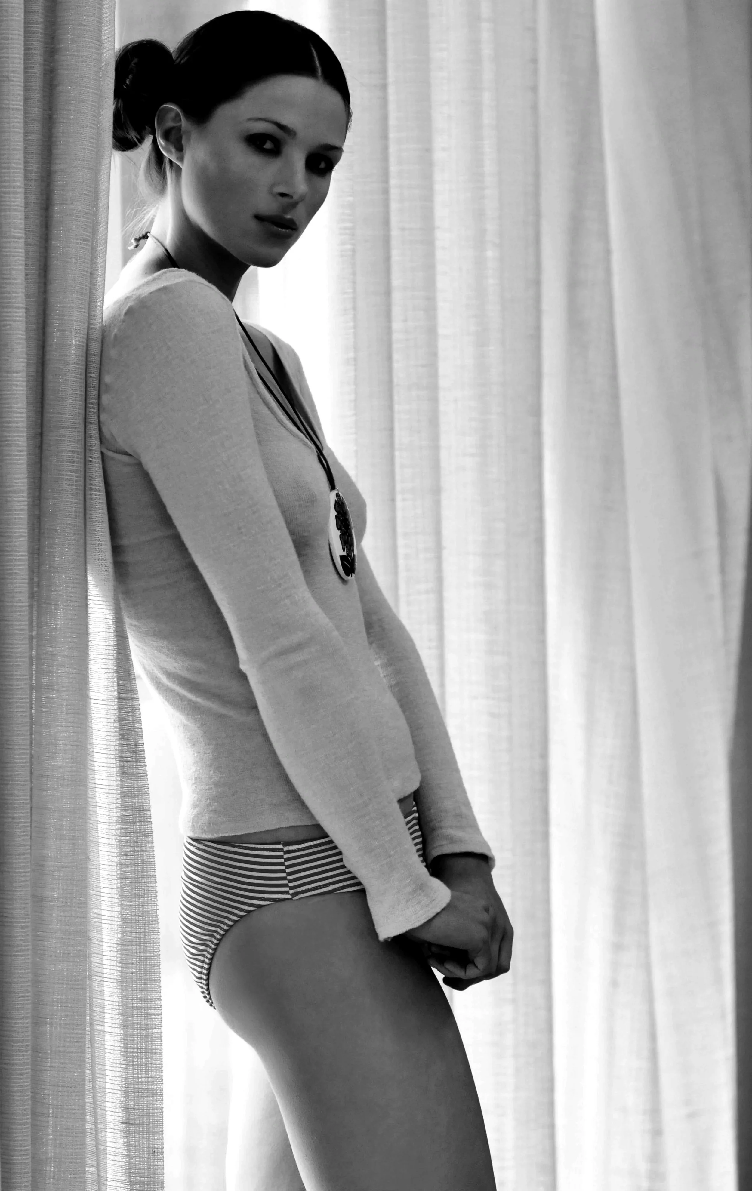 Black and white photo of a young woman standing next to curtains, wearing striped underwear, a long-sleeve top, and a necklace with a pendant, looking at the camera.