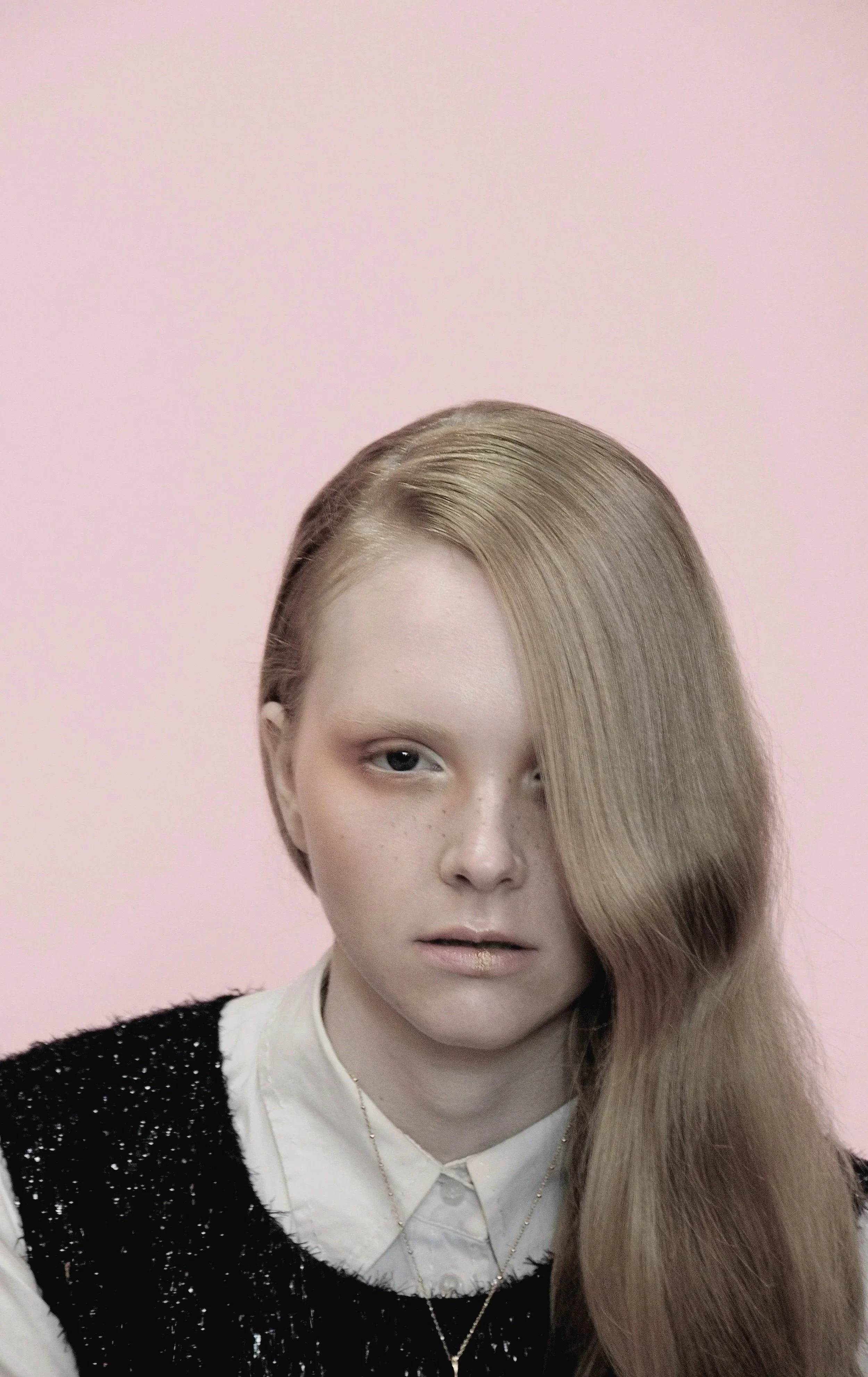 Close-up portrait of a young woman with long, wavy blonde hair partially covering one eye, wearing a white collared shirt under a black sequined vest, against a light pink background.