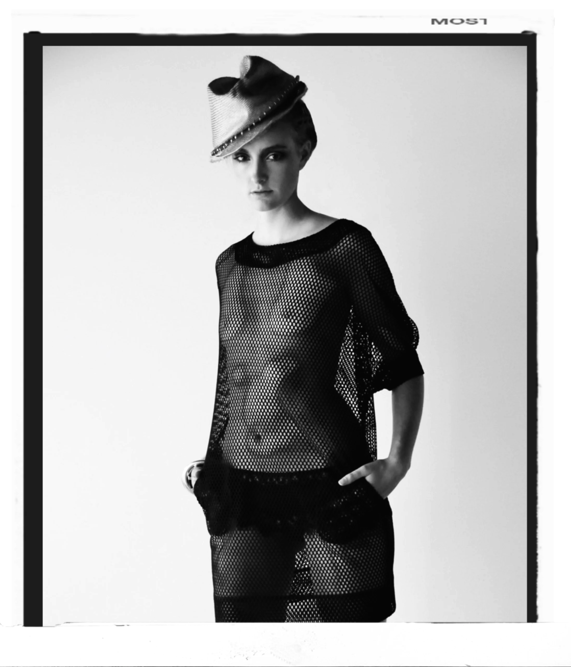 Black and white photograph of a woman wearing a sheer, see-through black dress and a fashion hat with a feather accent, standing against a plain background.