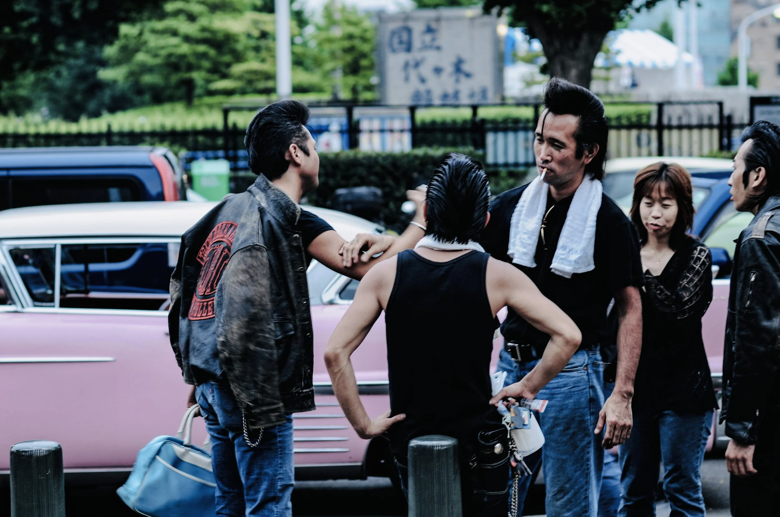 A group of people gathered outdoors near parked cars, engaged in conversation, with a green park and a sign in the background.