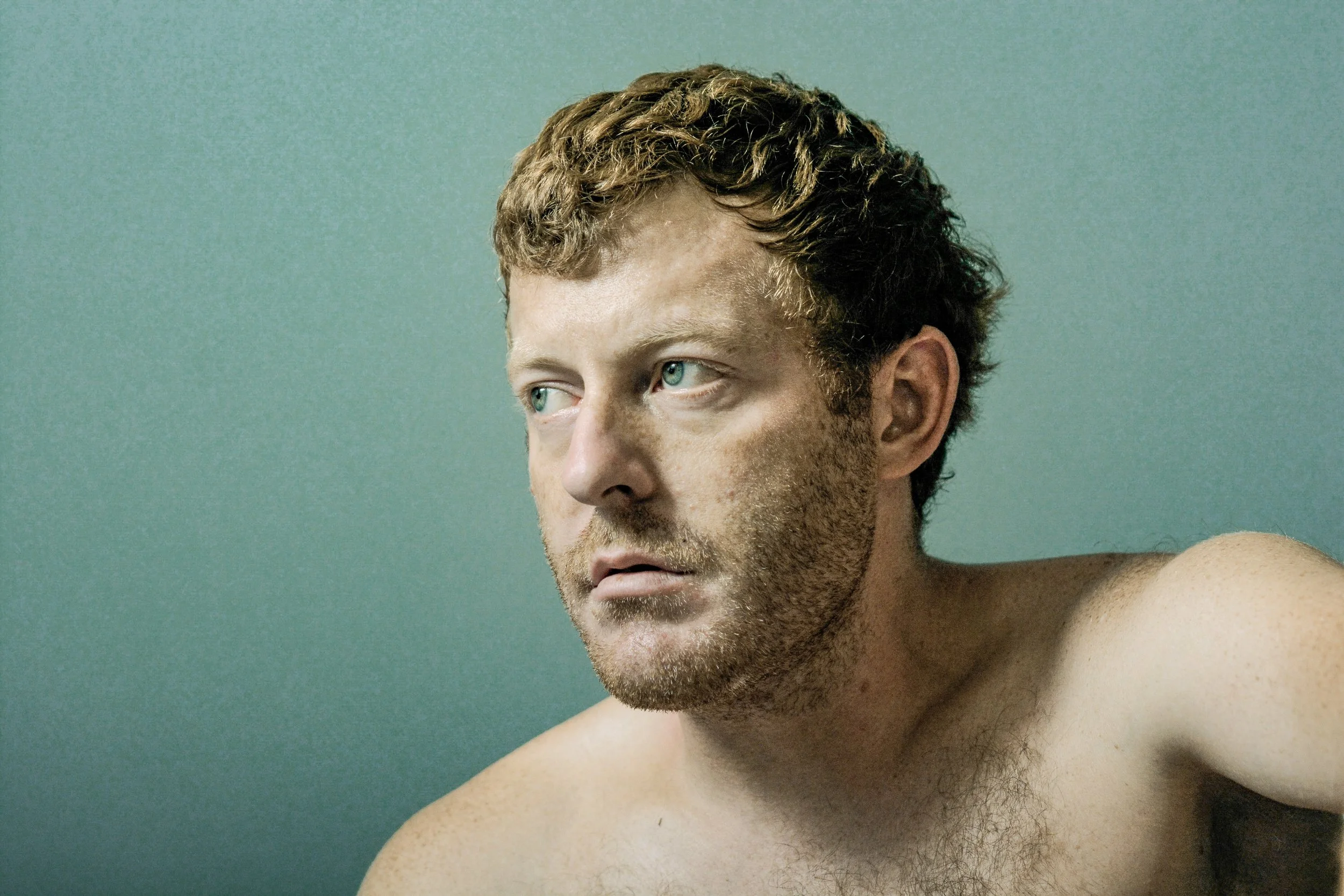 Close-up of a shirtless young man with curly, light brown hair and blue eyes, looking to the right, against a plain green background.
