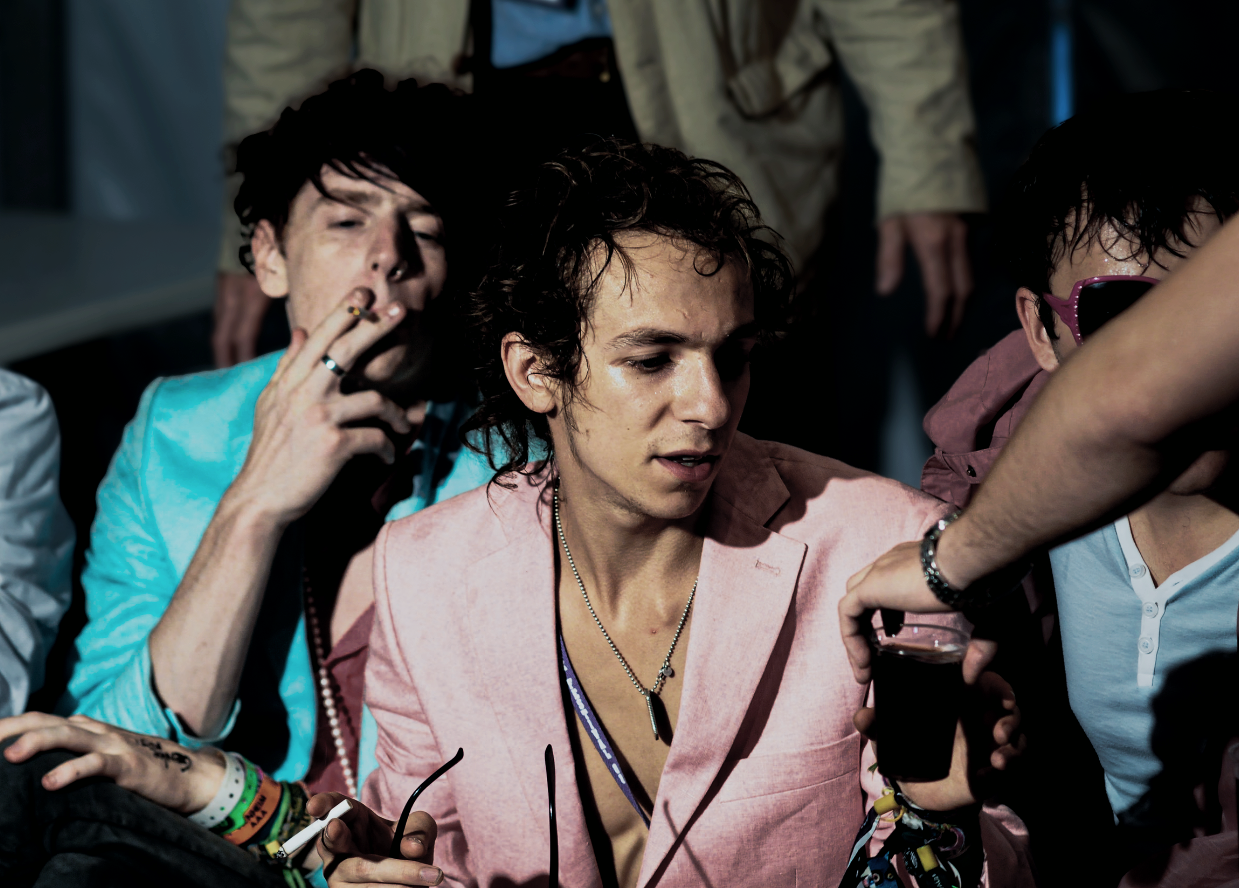 Three young men sitting closely together, engaged in conversation or activity, with one holding a cup of dark beverage, in a lively and casual setting.