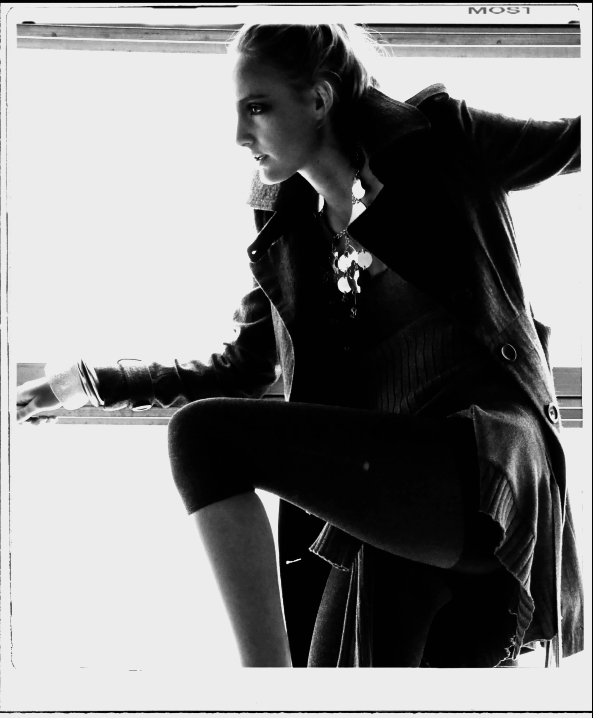 Black and white photo of a woman sitting by a window, wearing a dark jacket, scarf, and layered clothing, with her hair pulled back, looking outside.