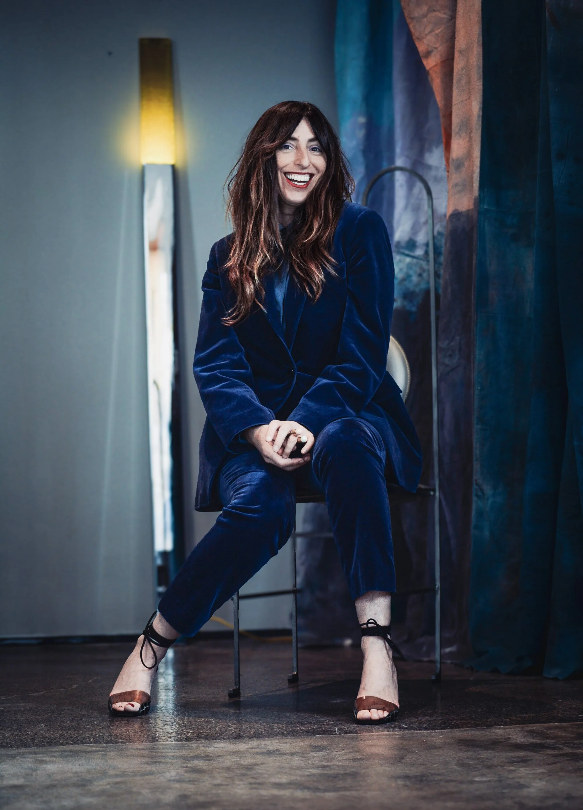 A woman with long brown hair, wearing a dark blue velvet blazer and pants, is sitting on a chair. She is smiling broadly and showing her teeth, with one arm resting on her knee and the other hand holding a smartphone. She is dressed in high heel shoe