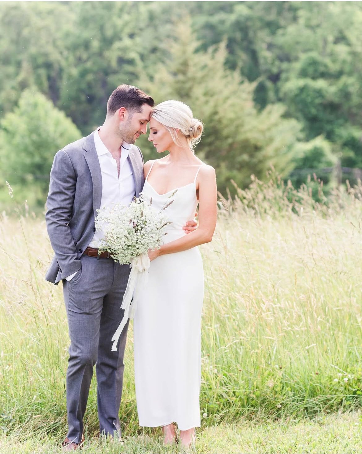 A bride and groom standing close in a grassy field, with heads touching and eyes closed, during their wedding. The bride is holding a bouquet of white flowers, and the groom is wearing a gray suit with an open-collared shirt. The background features