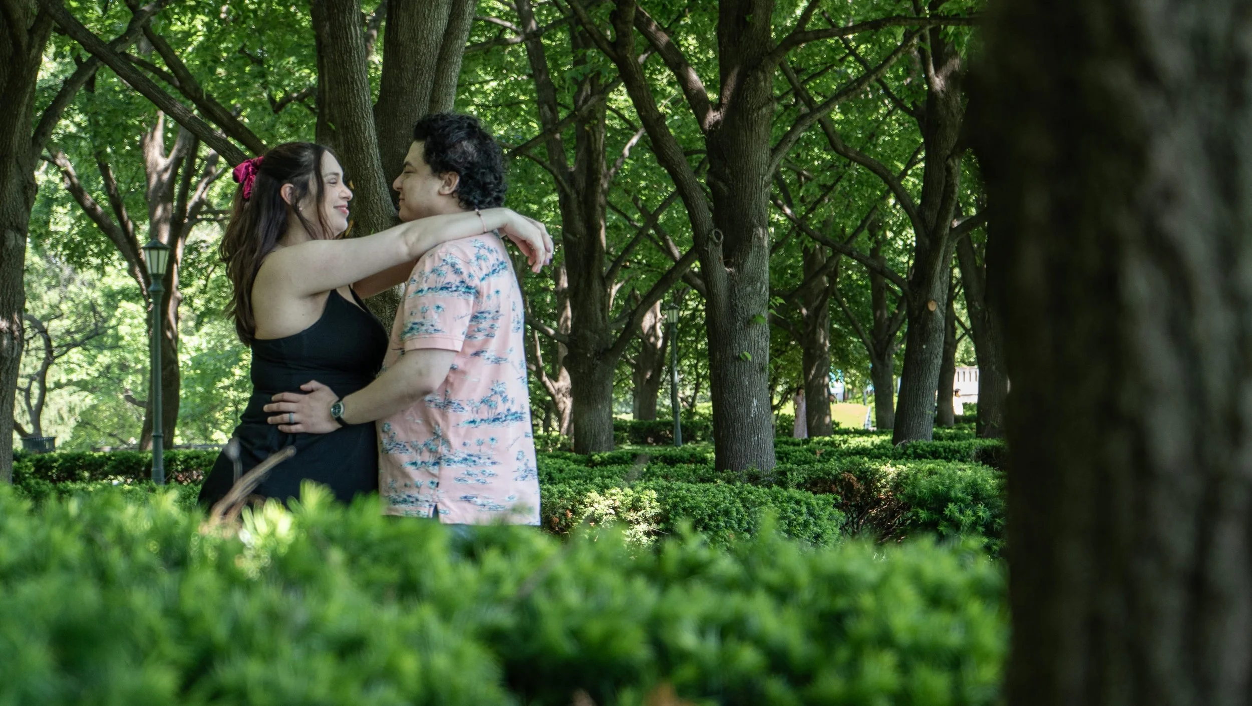 A couple is standing close together in a park with green trees and bushes, embracing and looking into each other's eyes.