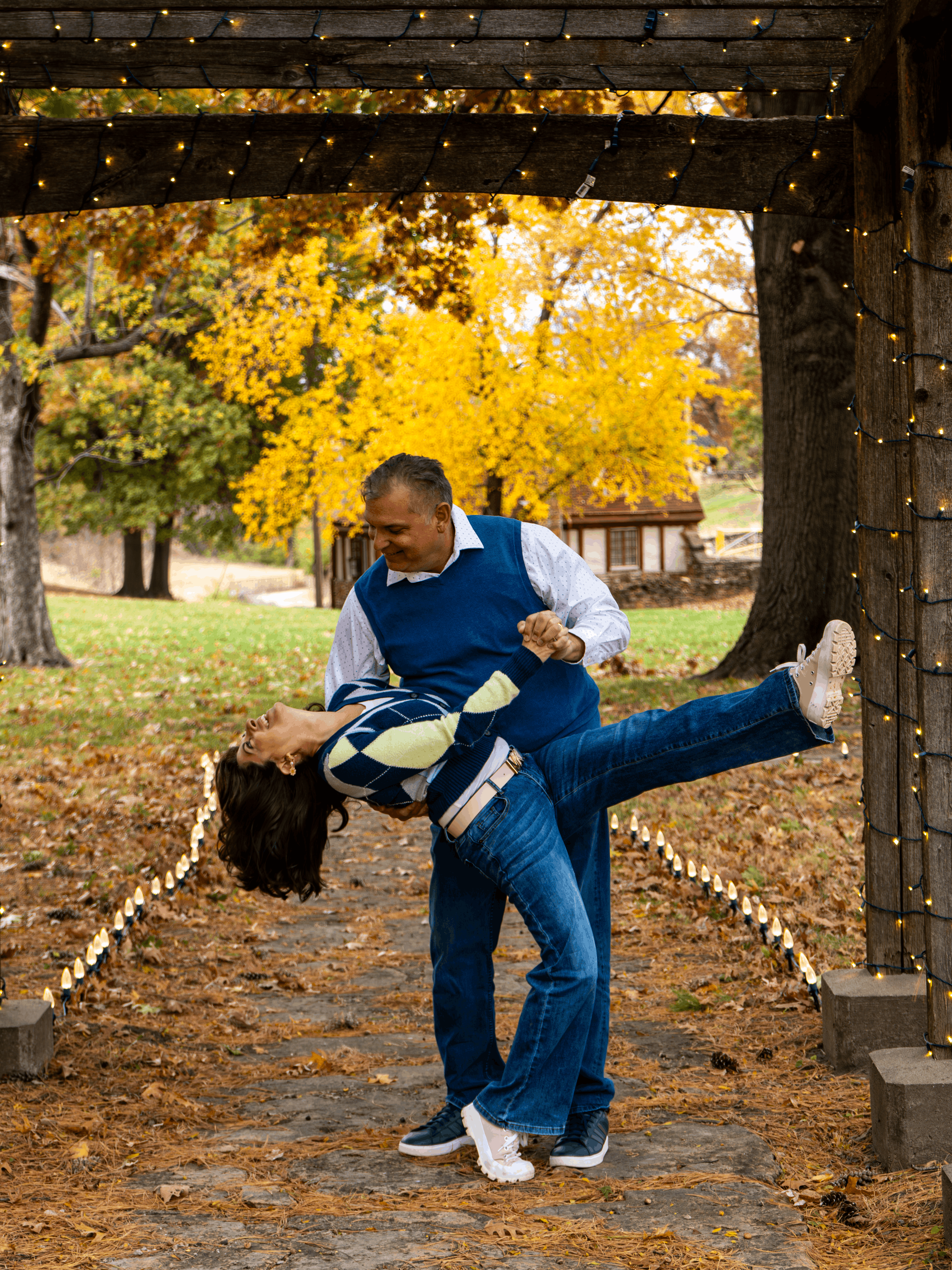 A man giving a piggyback ride to a young girl in an outdoor park with autumn trees, fallen leaves, and Christmas lights in the background.