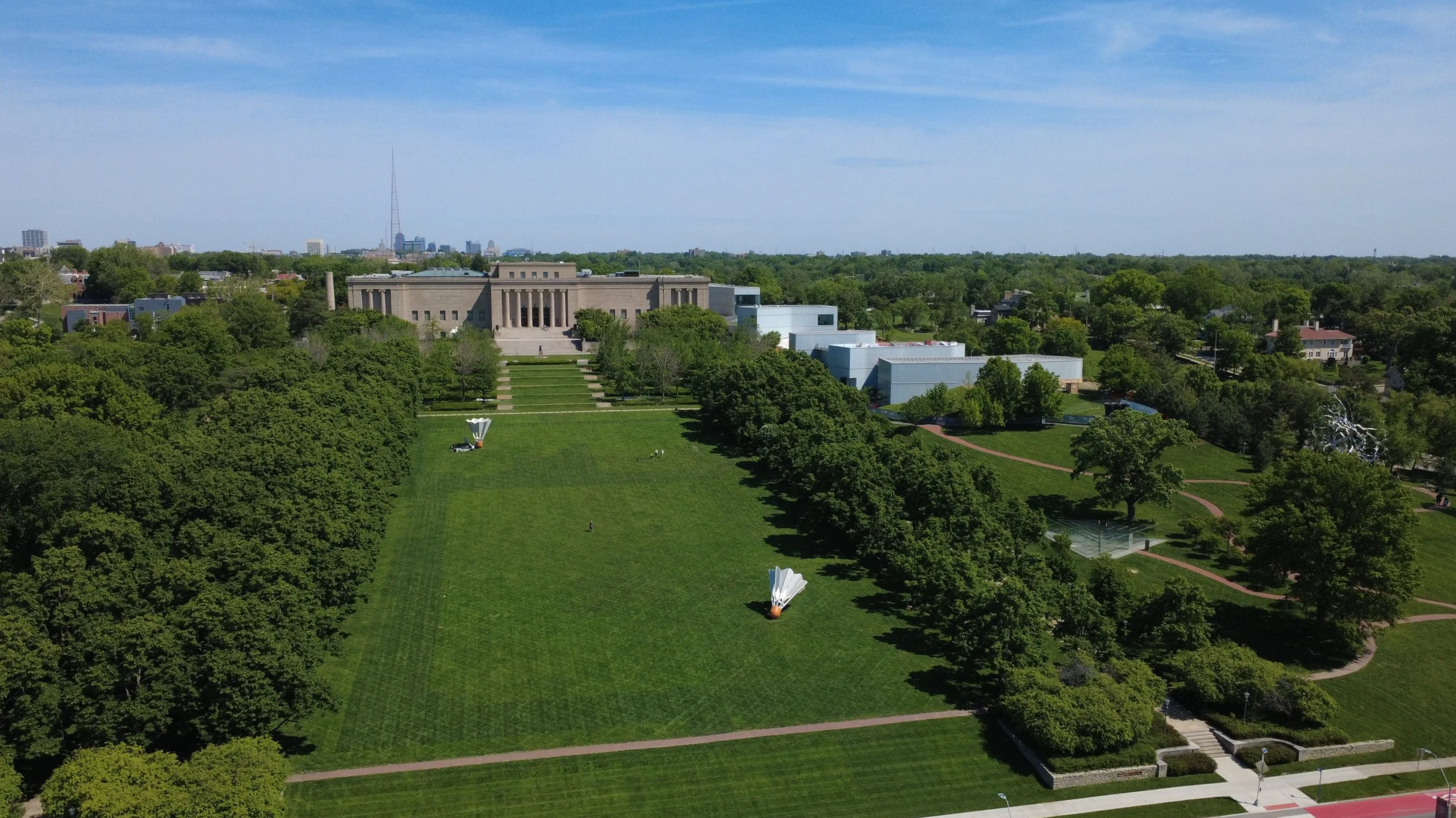 Aerial view of a large park with green lawns, trees, pathways, and a prominent building with classical architecture in the background, under a blue sky.