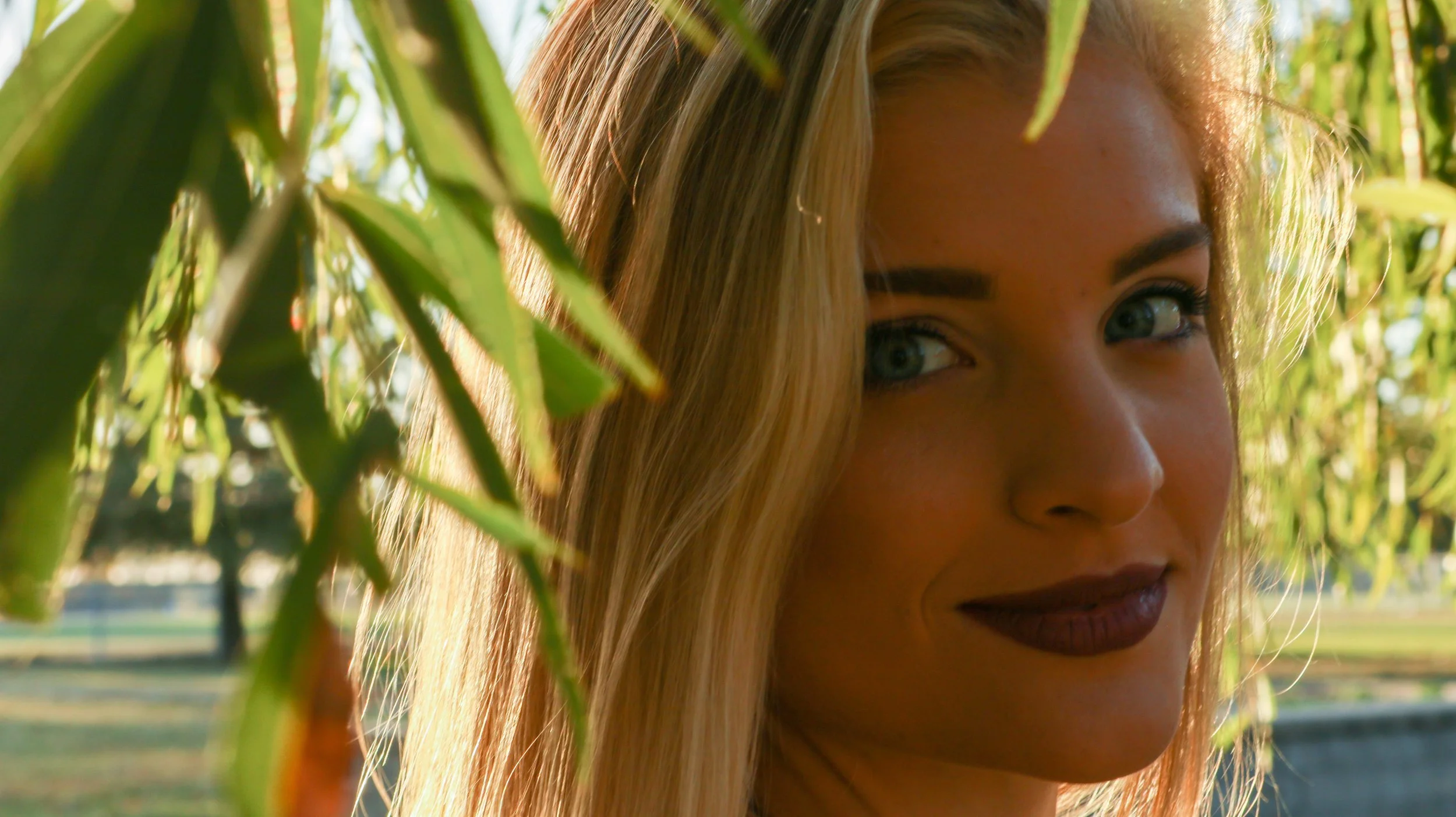 Part of a young woman's face with blonde hair and green eyes peeking through green foliage outdoors during daytime.