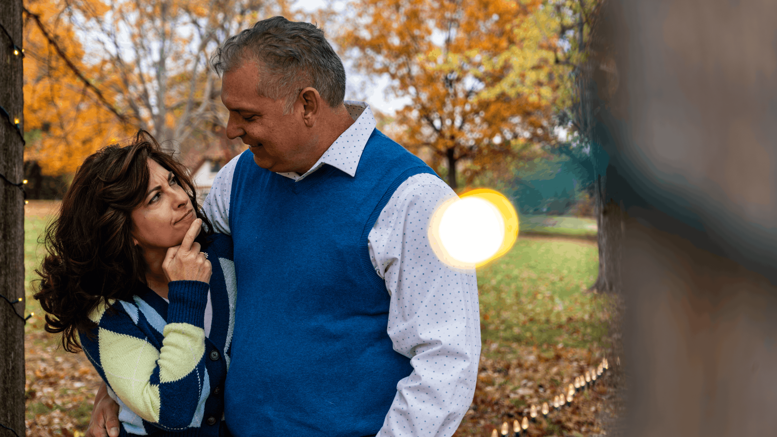 A man and woman sharing an intimate moment outdoors during fall, with orange leaves on trees in the background, and sunlight creating lens flare.