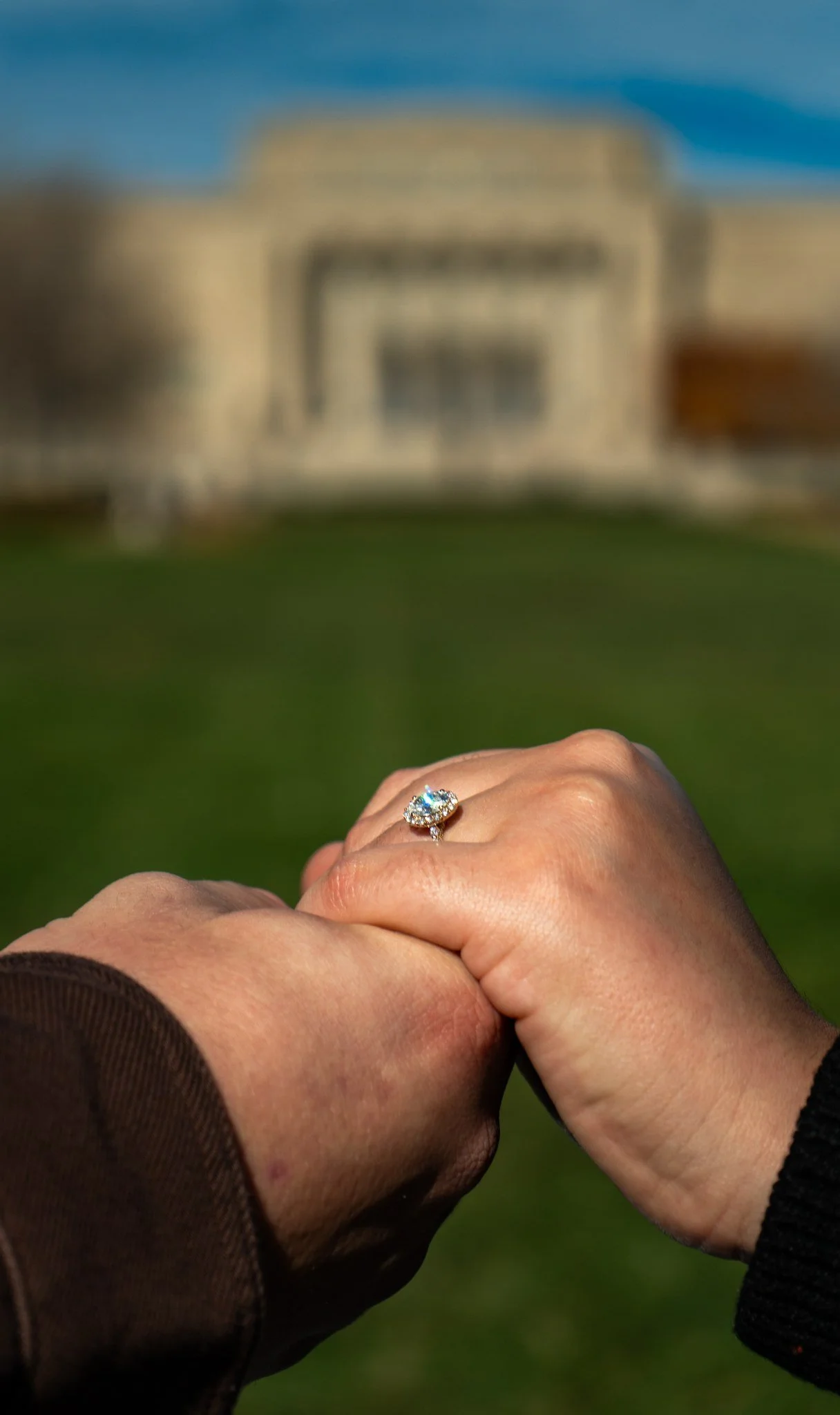 Two hands clasped together, one with a diamond engagement ring, against a blurred background of a building and clear blue sky.