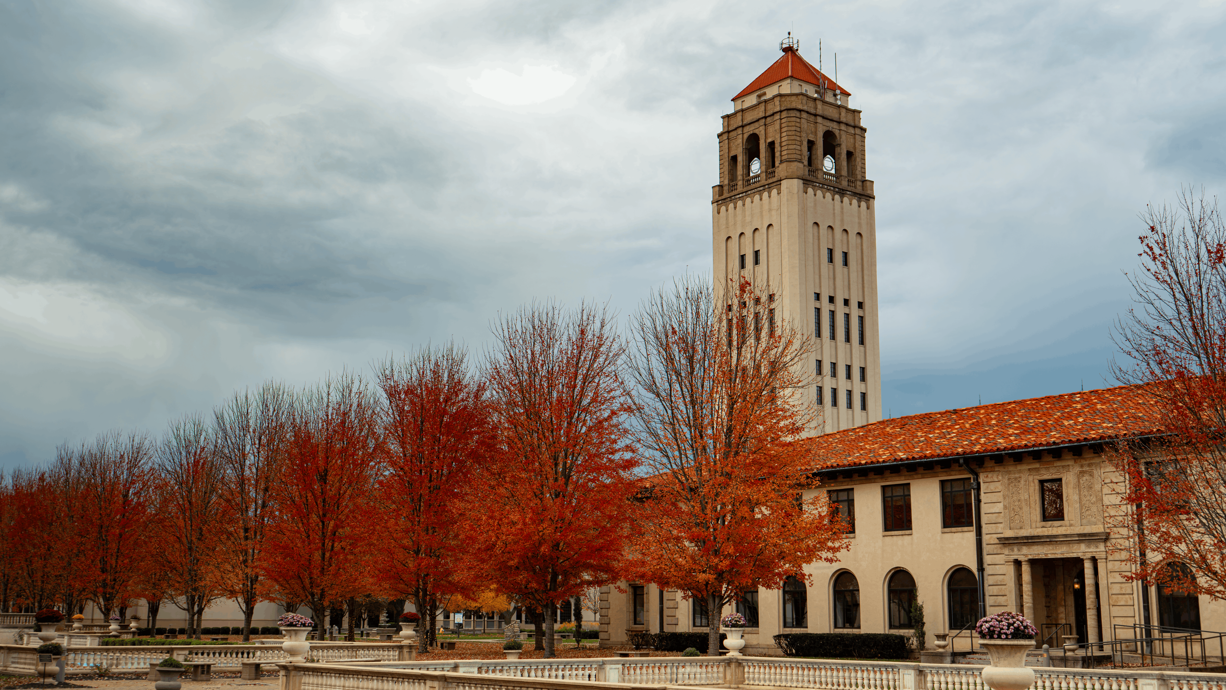 A tall clock tower with a red roof stands against a cloudy sky, surrounded by autumn trees with orange and red leaves. A building with arched windows and decorative stonework is visible in the foreground.