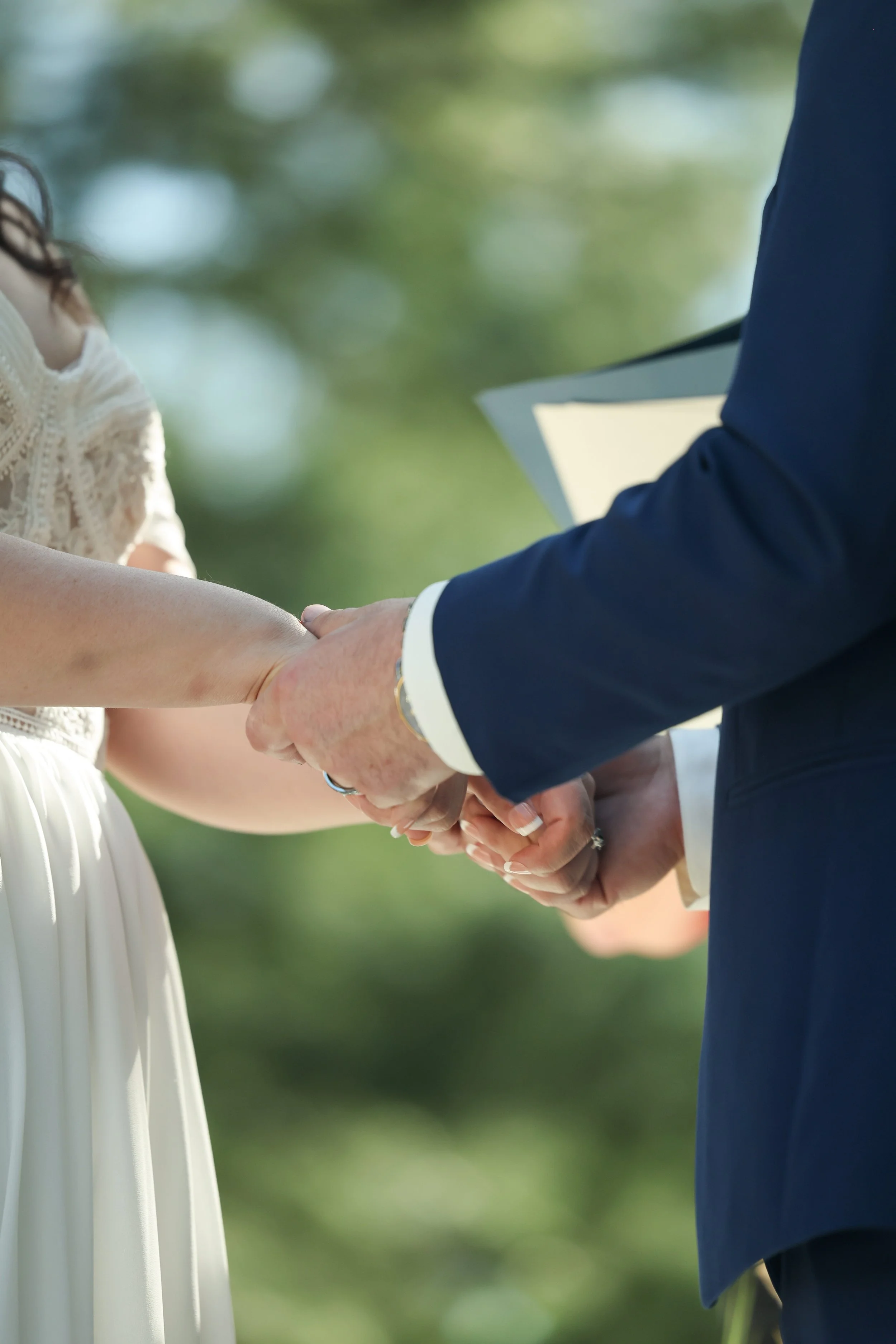 A couple holding hands during a wedding ceremony, with the bride wearing a white dress and the groom in a dark suit, outdoors with blurred green foliage in the background.