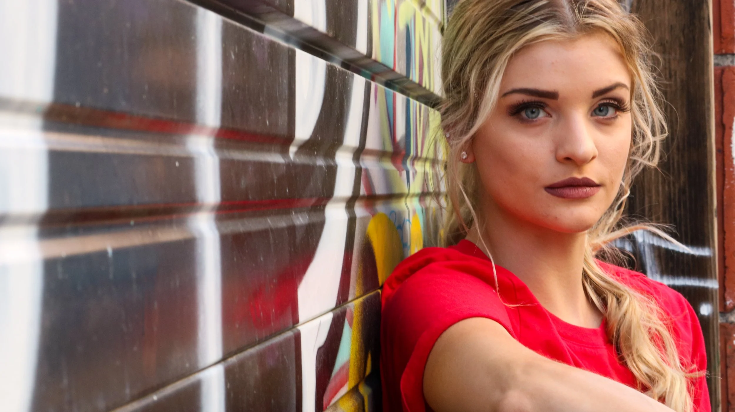 A young woman with blonde hair and blue eyes wearing a red shirt, leaning against a colorful graffiti-covered wall.